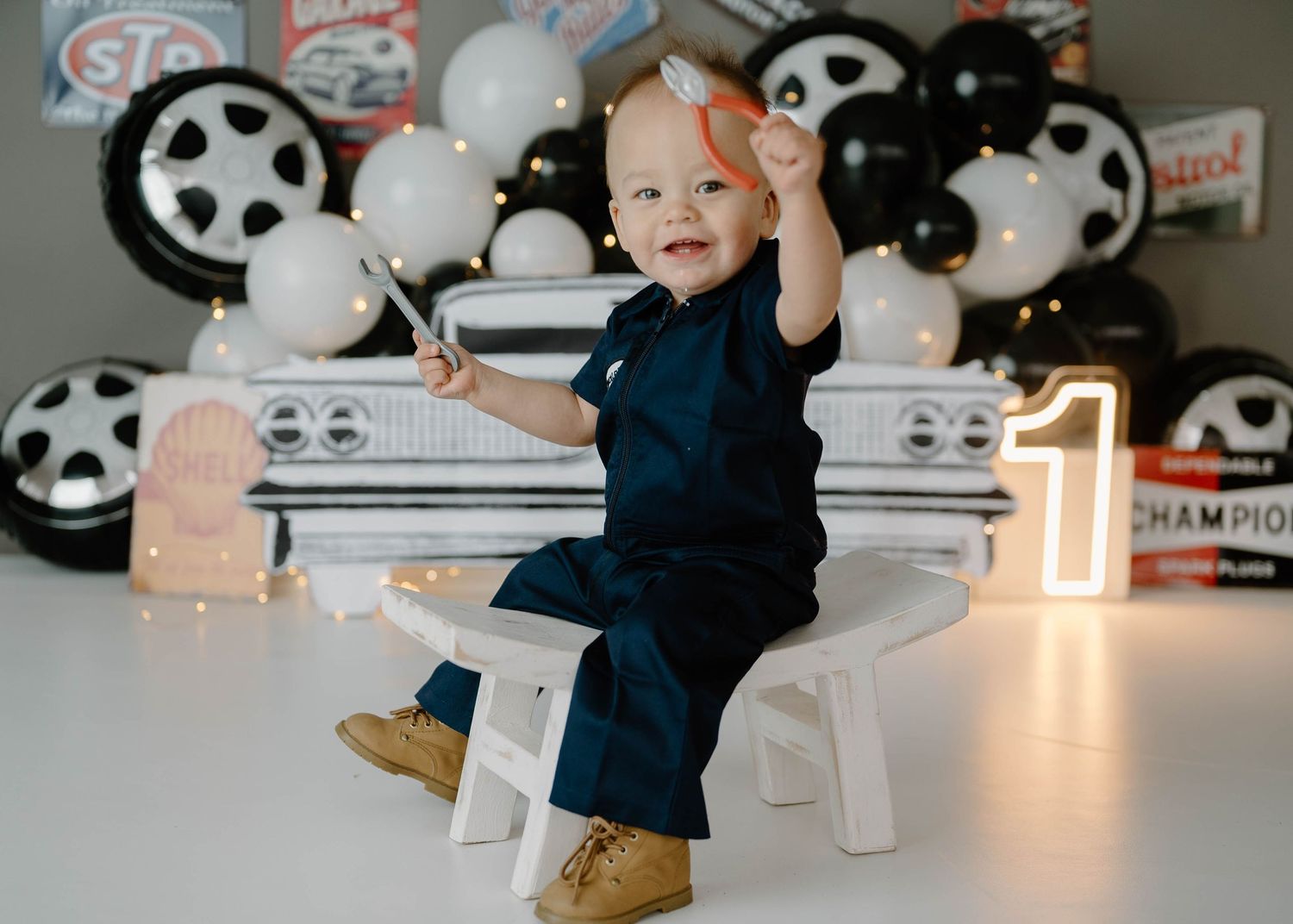 Baby boy in navy outfit sits on white chair at car-themed first birthday party with balloon backdrop and number 1.