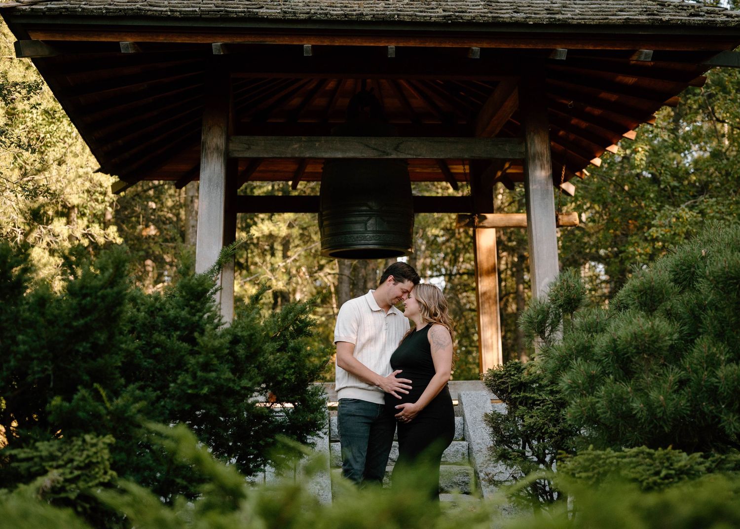 Expecting couple shares a romantic kiss beneath a wooden gazebo surrounded by lush garden greenery in Devon Alberta.
