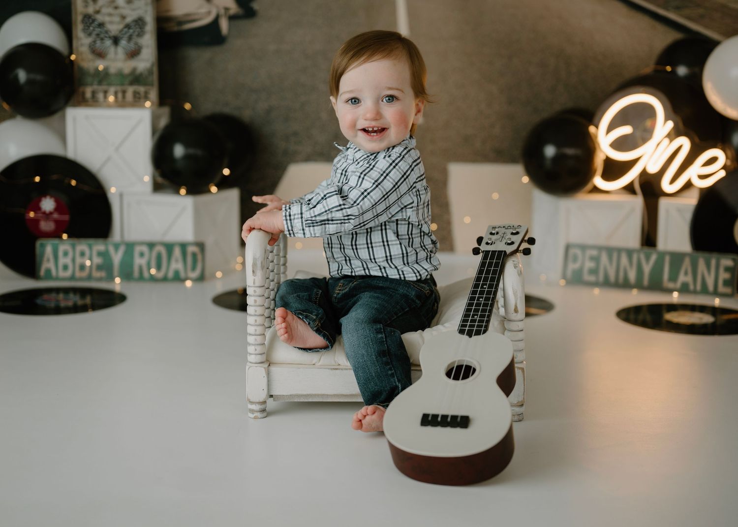 Smiling toddler boy in plaid shirt sits on white chair with Beatles-themed Abbey Road birthday decorations.