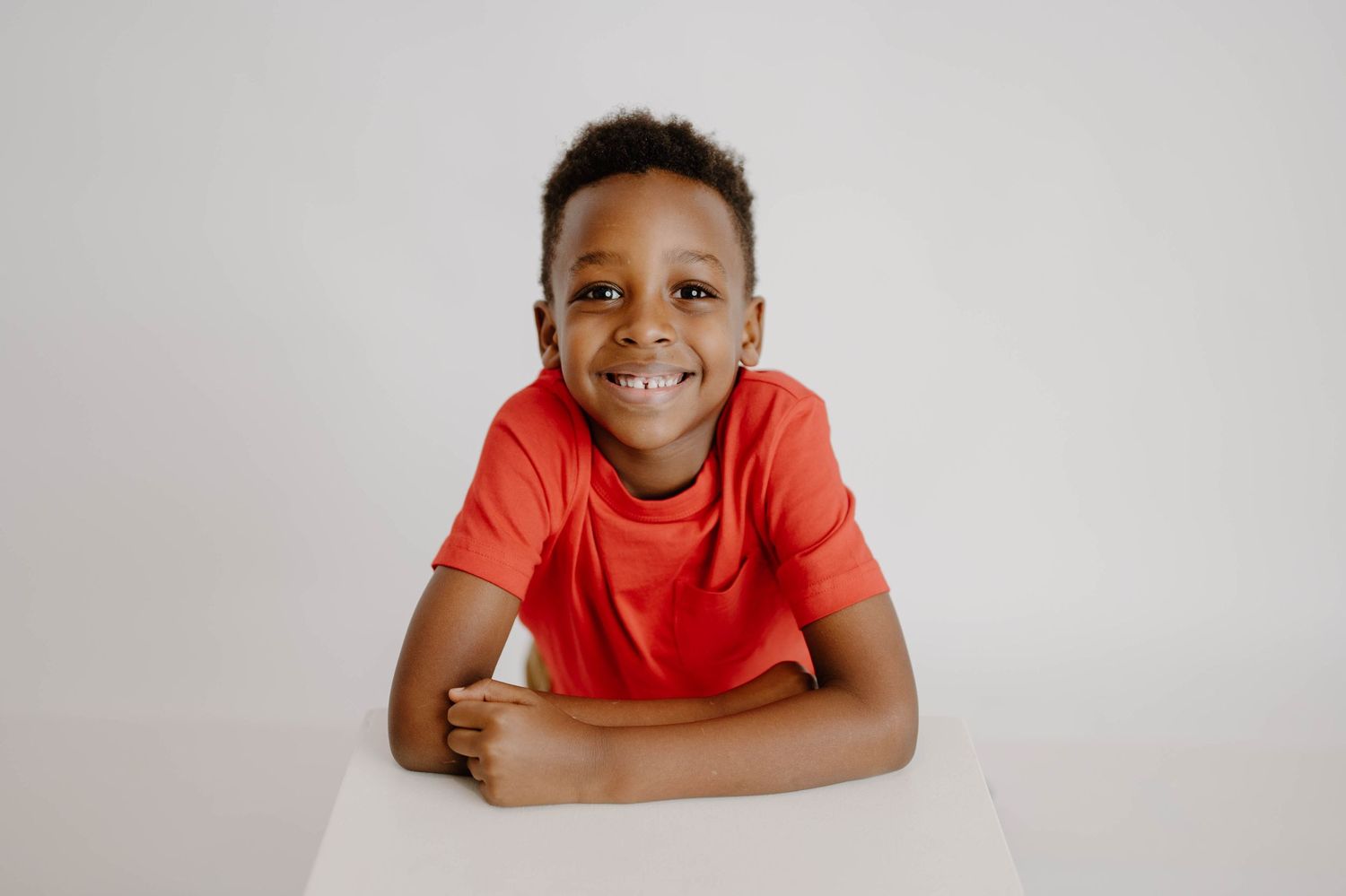 Happy young Black boy in a red t-shirt sitting at a white table smiling at the camera.