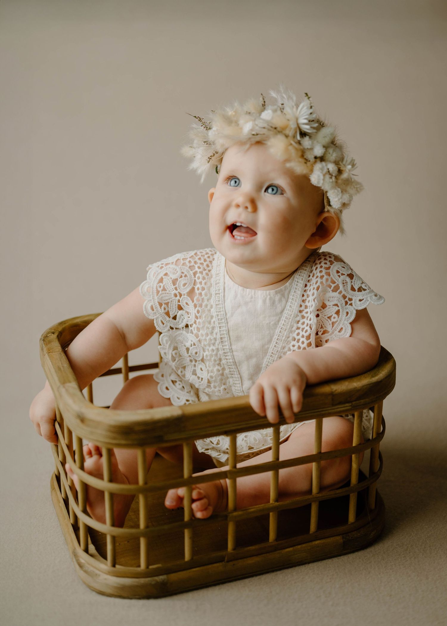 Adorable baby girl wearing a floral crown and lace romper sitting in a wicker basket for milestone photos.