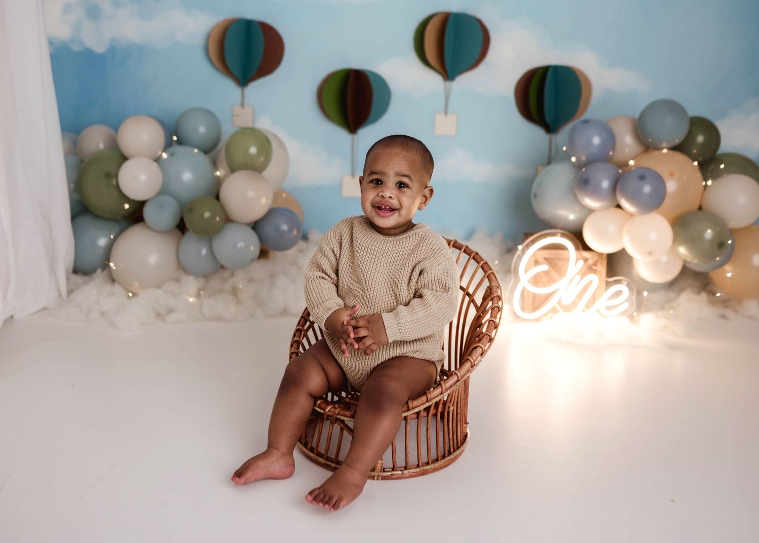 Smiling baby boy sitting in a rattan chair at a hot air balloon themed first birthday photo session with neon One sign.