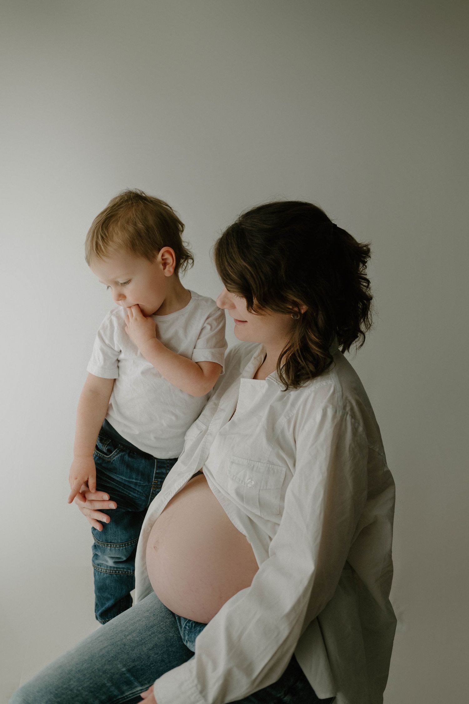 Pregnant mom in open white shirt sits with toddler son beside her exposed belly in soft light.