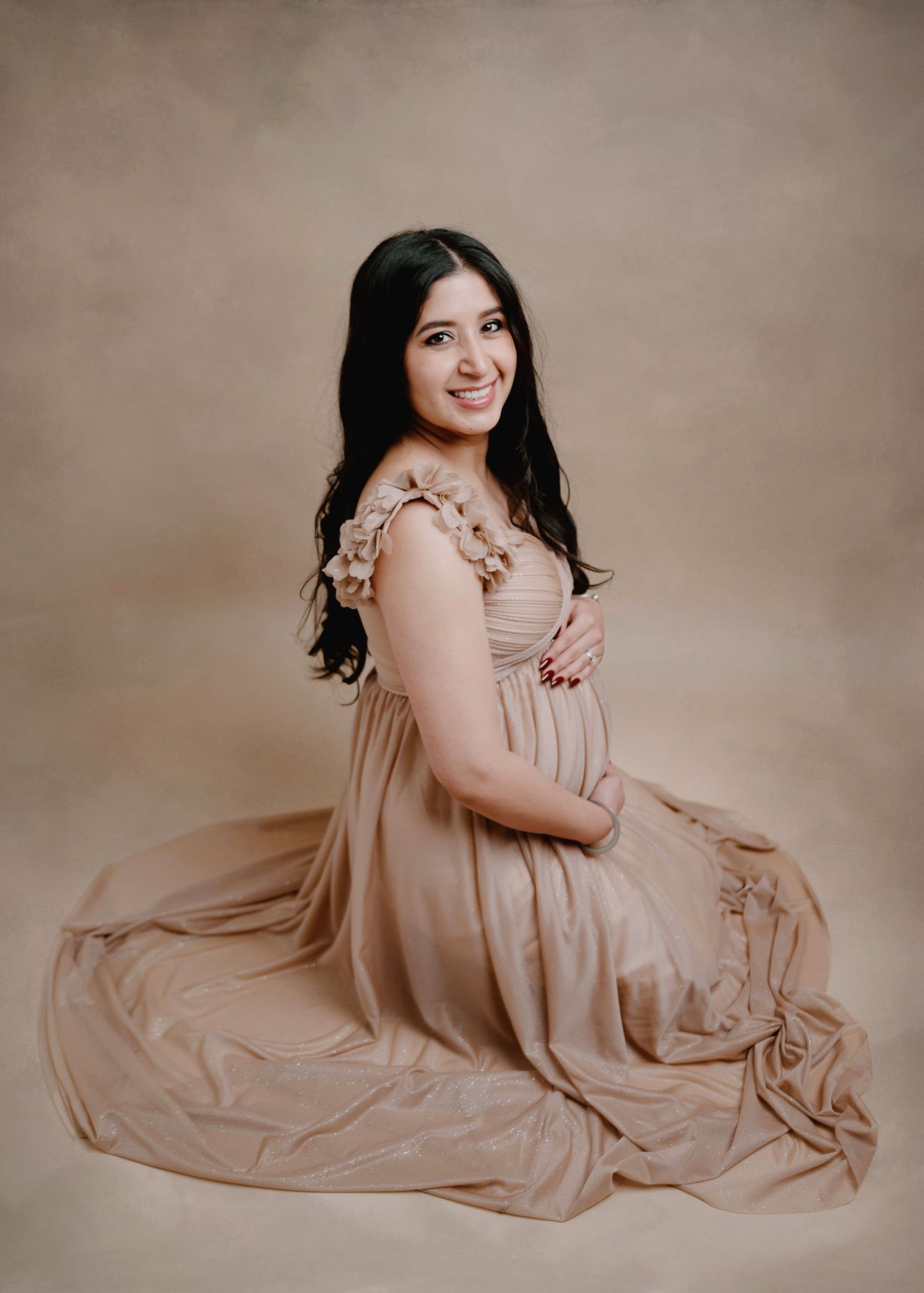 A pregnant woman in a golden beige gown sits gracefully on the floor in a studio maternity session.