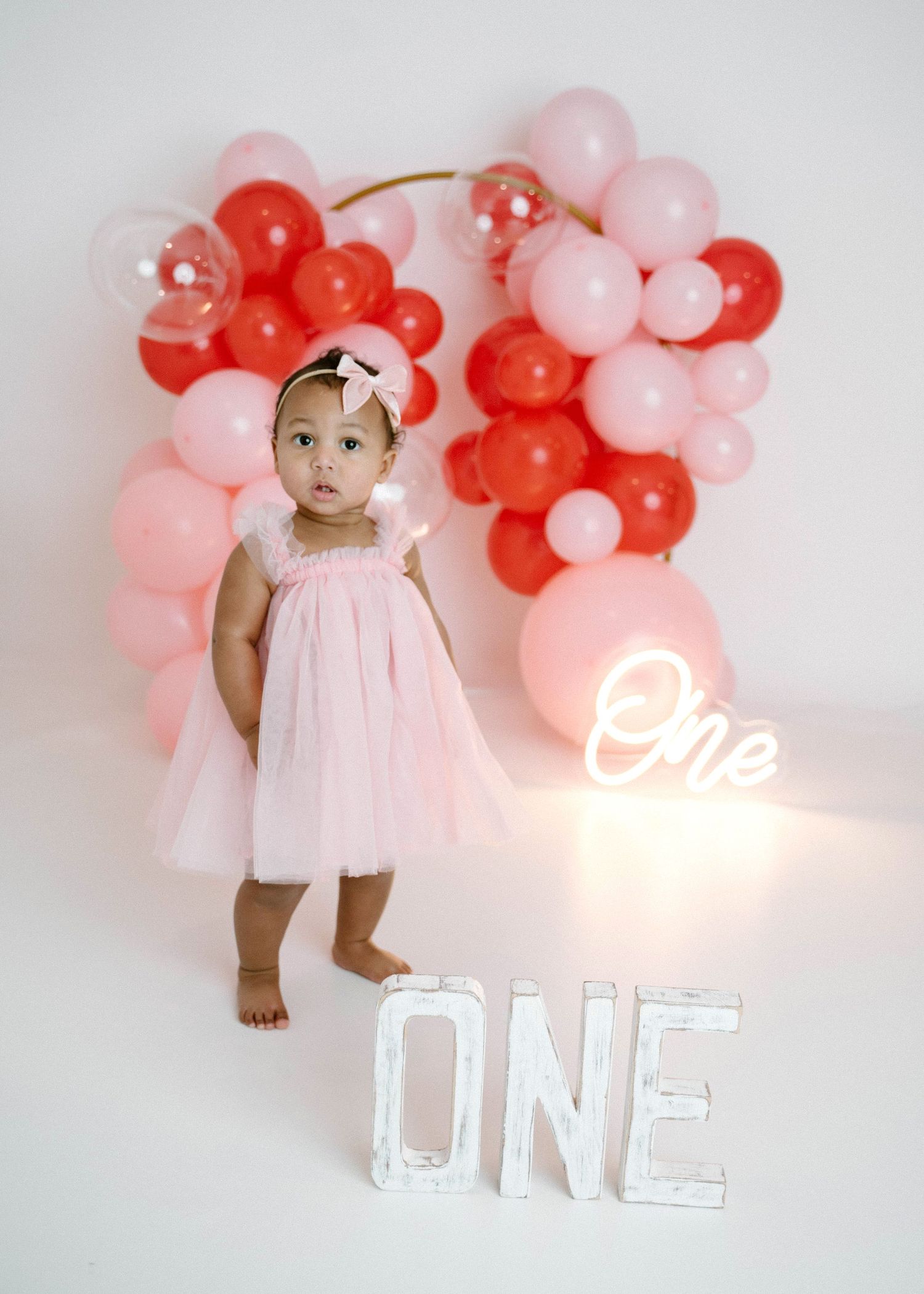 Baby girl in pink dress stands beside ONE letter sign with heart-shaped red pink balloon wreath.