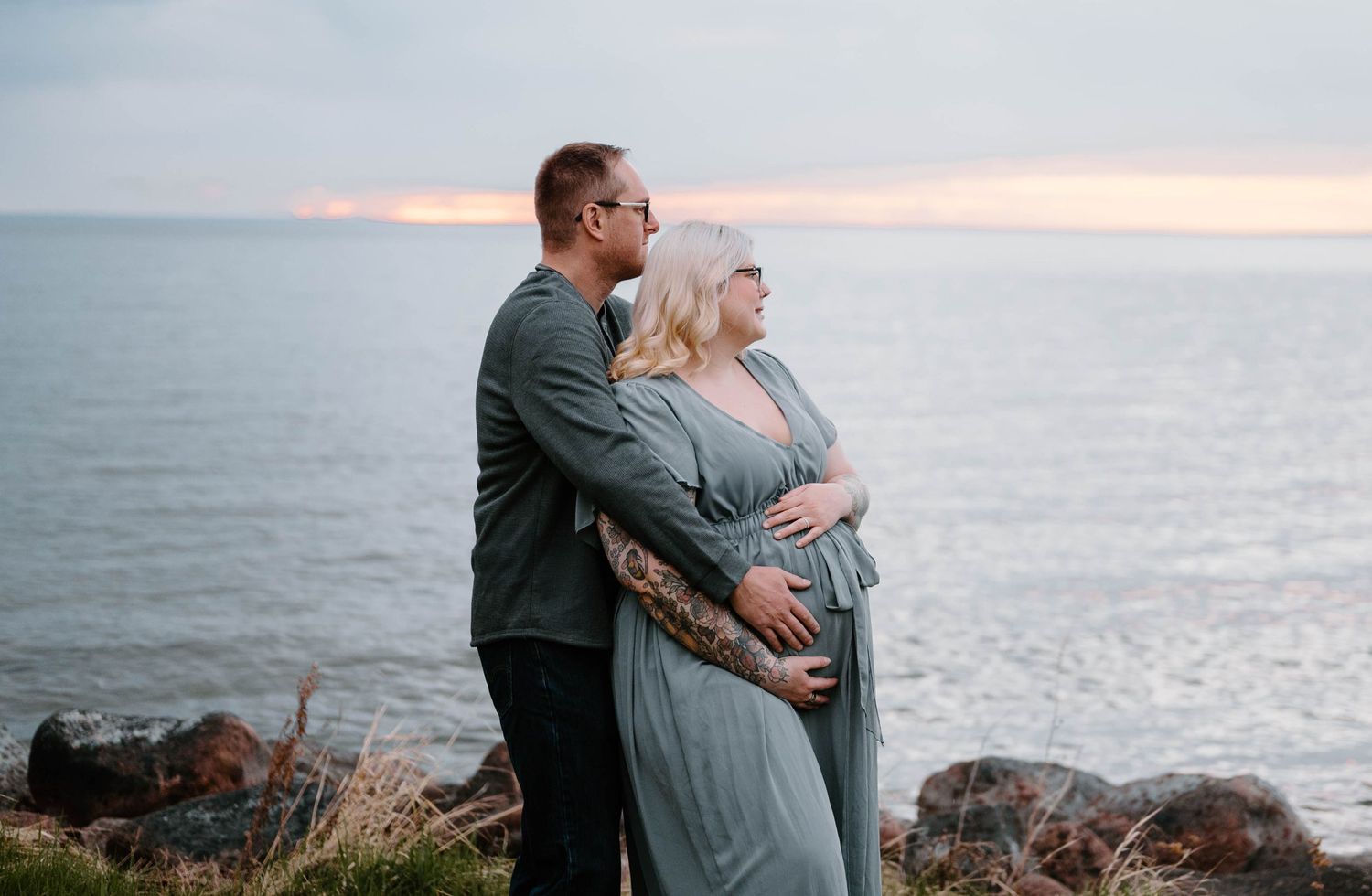 Couple embracing at ocean cliff during sunset, pregnant woman in grey dress with partner in green jacket.