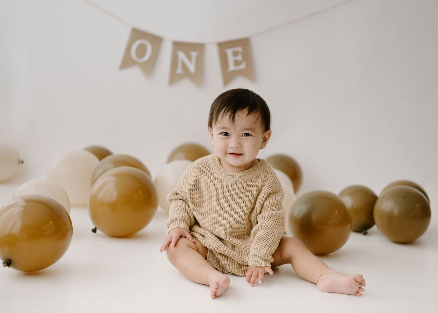 A smiling baby in a beige knit outfit sits among gold balloons with an ONE banner for a first birthday cake smash photo.