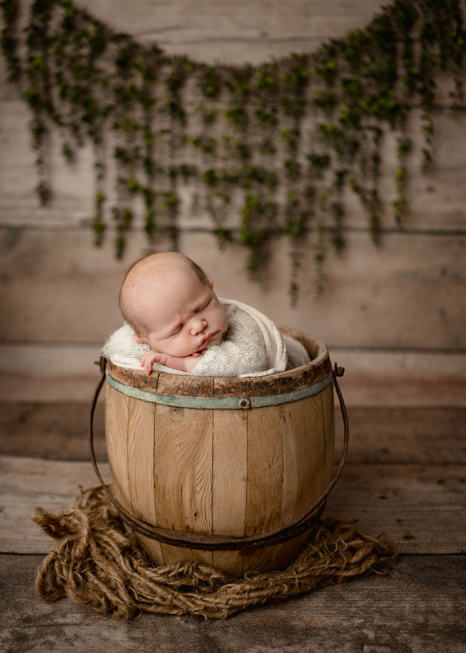 Sleeping newborn baby nestled in a rustic wooden bucket with soft white wrap, surrounded by greenery backdrop in cozy leduc portrait studio.