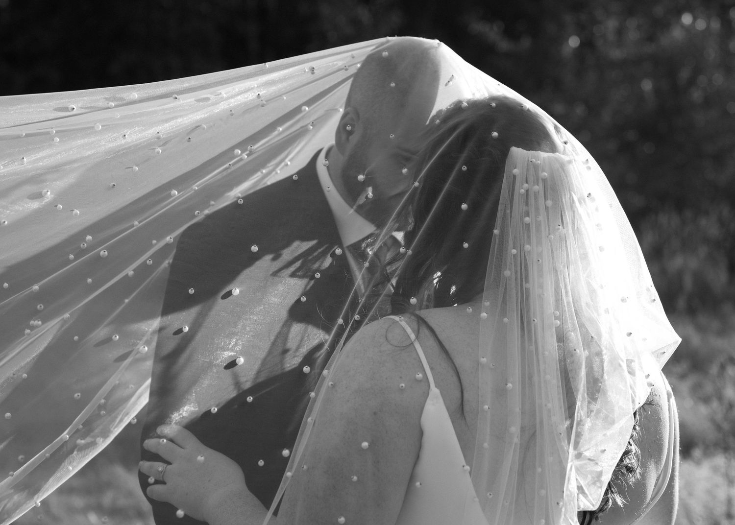 Black and white photo of bride and groom kissing beneath a pearl-dotted wedding veil outdoors.