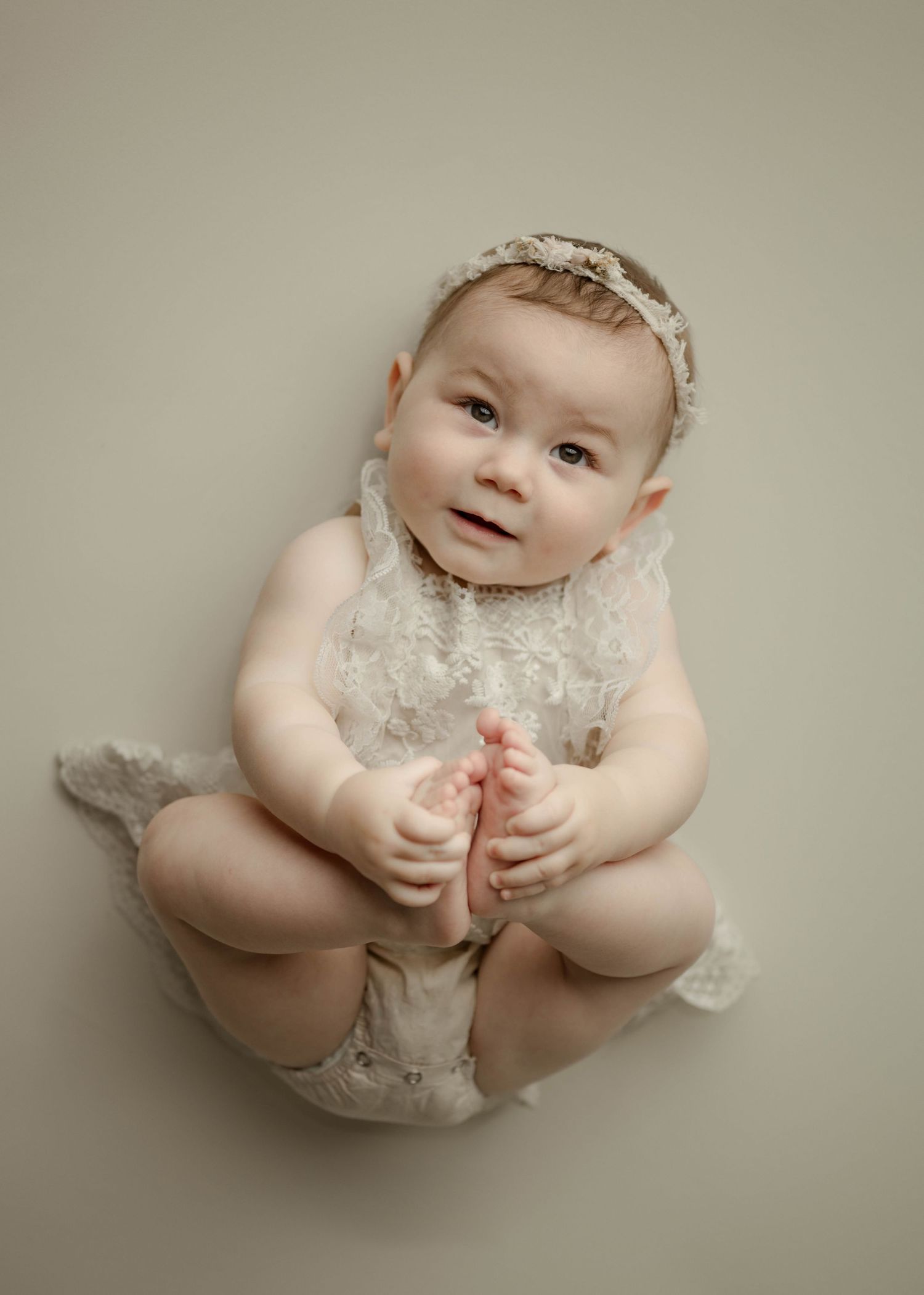Cute baby girl in lace romper and bow headband holding her feet during professional studio portrait photography session.
