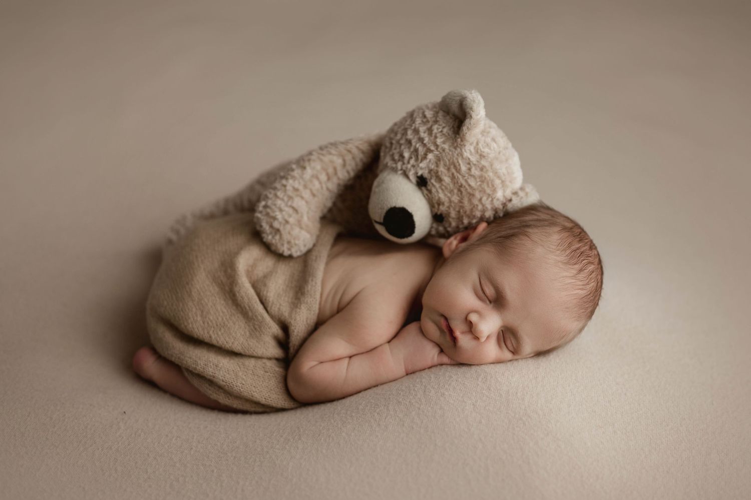Sleeping newborn baby in beige wrap posed with small teddy bear on cream studio backdrop.