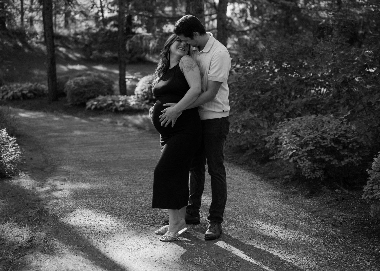 Couple embraces tenderly in black and white maternity photo on a tree-lined garden path in Botanical Gardens.