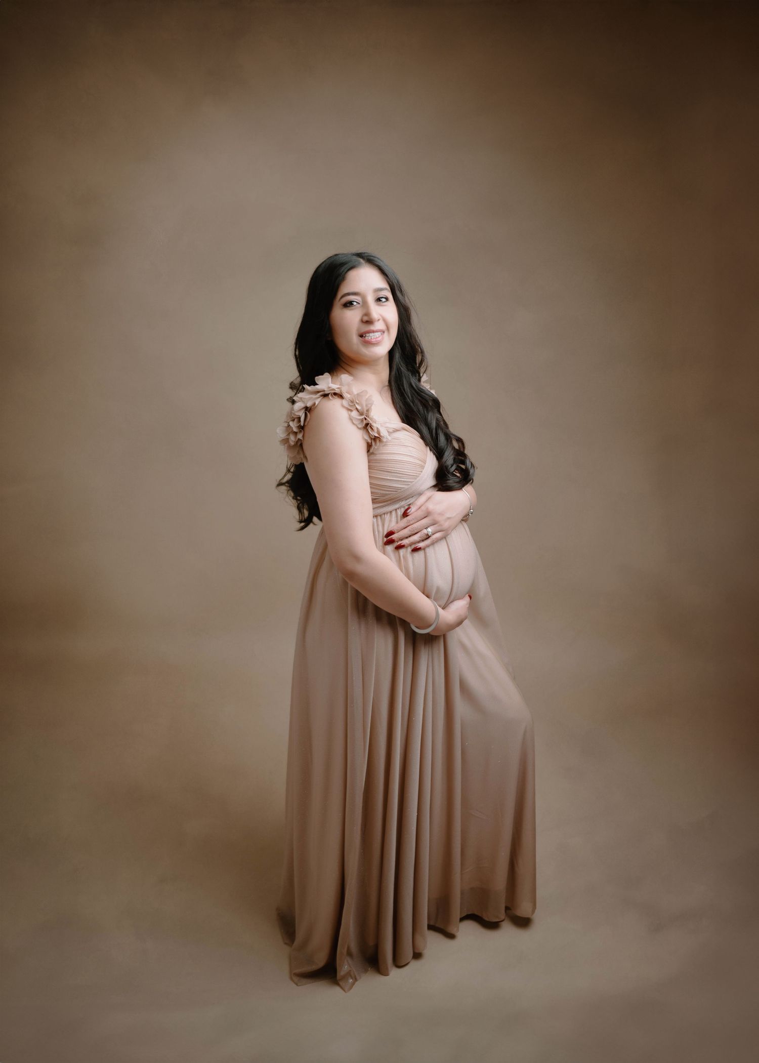 A smiling pregnant woman in a tan ruffled gown holds her baby bump in a Leduc studio maternity photo.