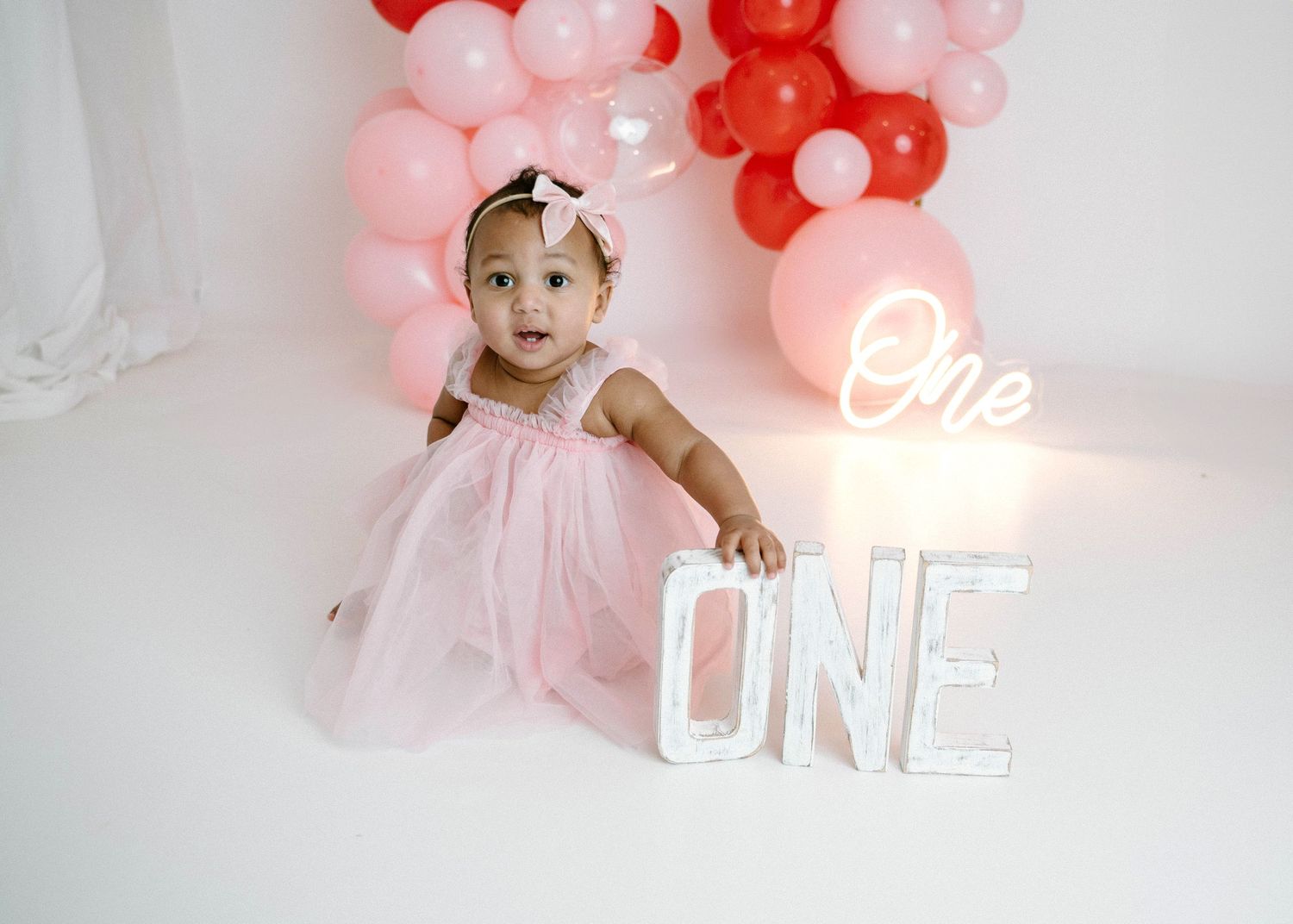 Baby girl in pink tulle dress poses with lit ONE letter sign and pink red balloon arch.
