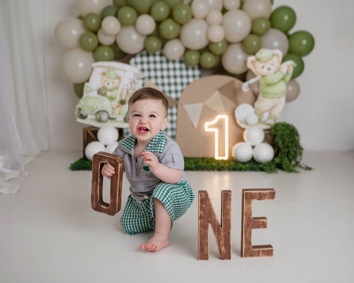 Happy baby boy poses with wooden ONE letters at golf-themed first birthday backdrop with green and white balloons.