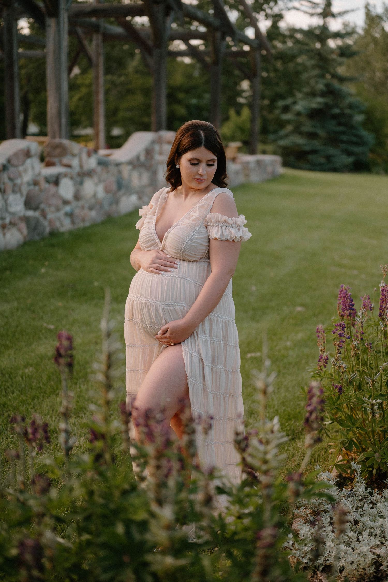 Pregnant woman in flowing lace dress cradles baby bump among wildflowers during golden hour maternity session.