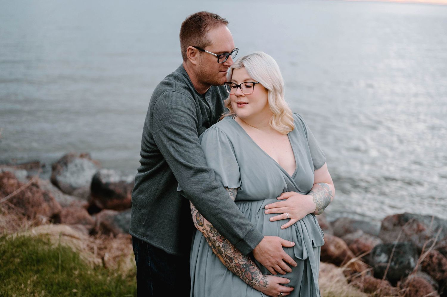 A couple embraces near rocky shoreline, man kisses woman's head while holding her baby bump in a tender maternity photo.