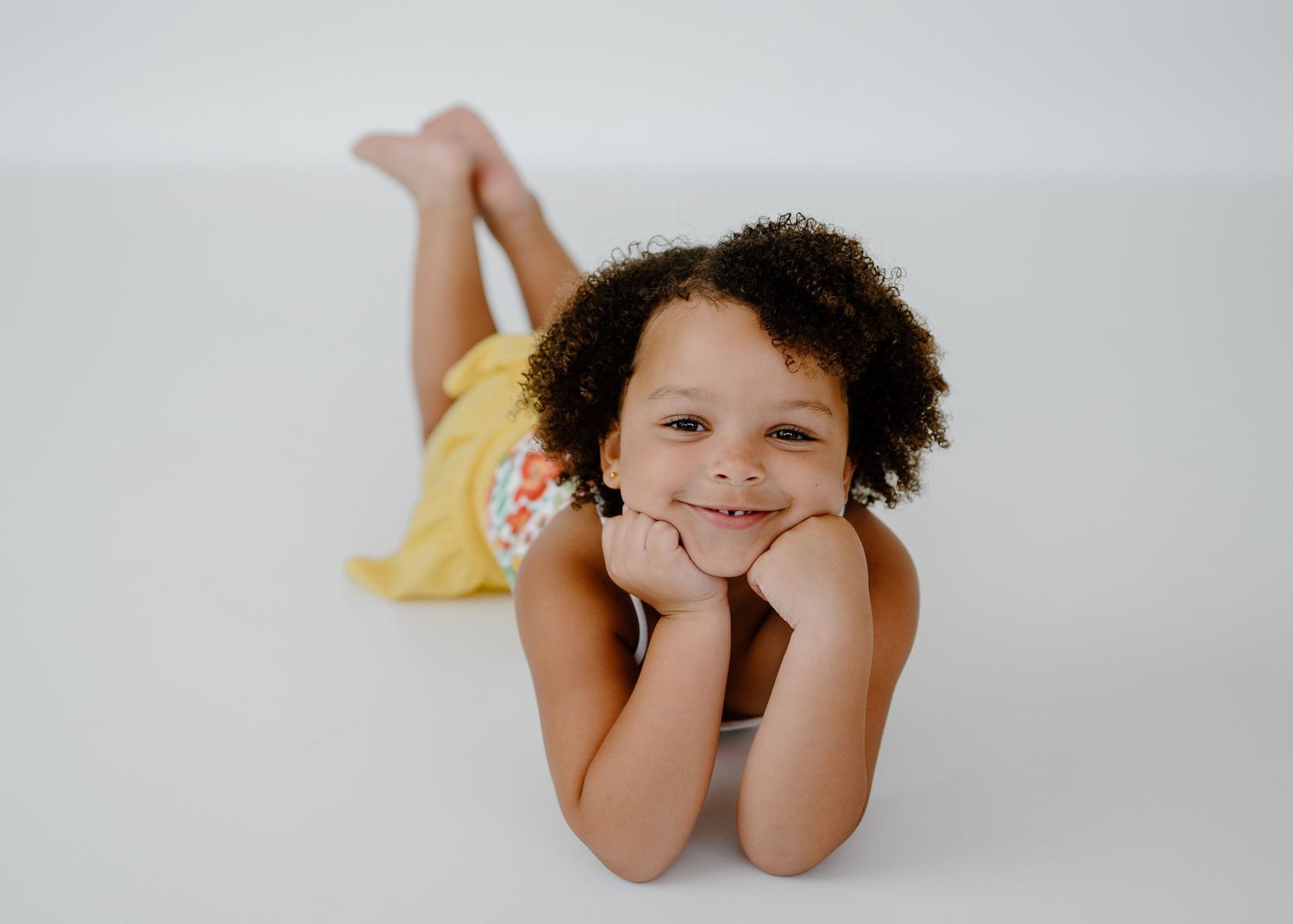 Smiling young girl with curly hair lying on white floor in yellow floral outfit.