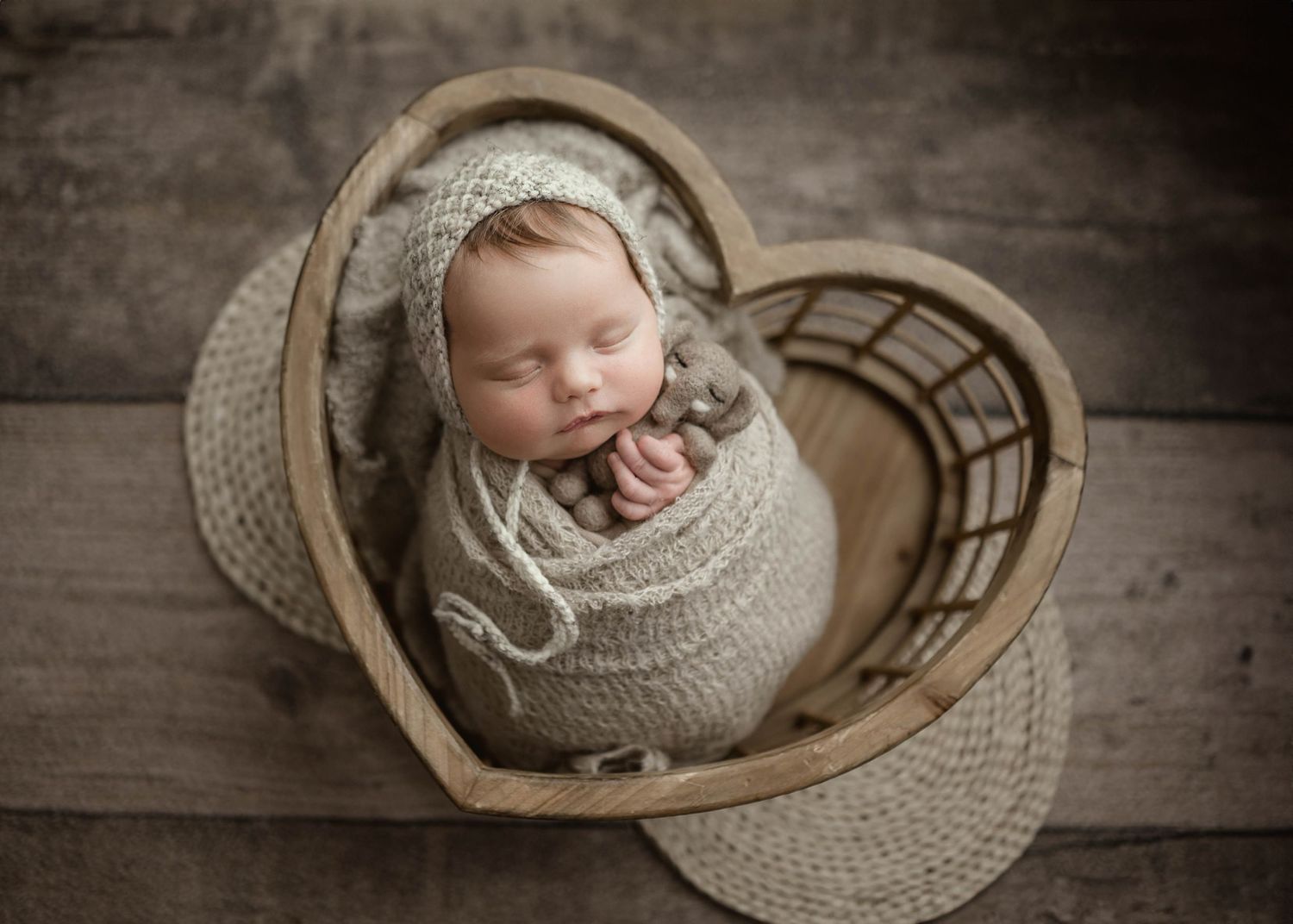 Newborn baby swaddled in gray knit wrap sleeping in a wooden heart-shaped basket for a studio photo.