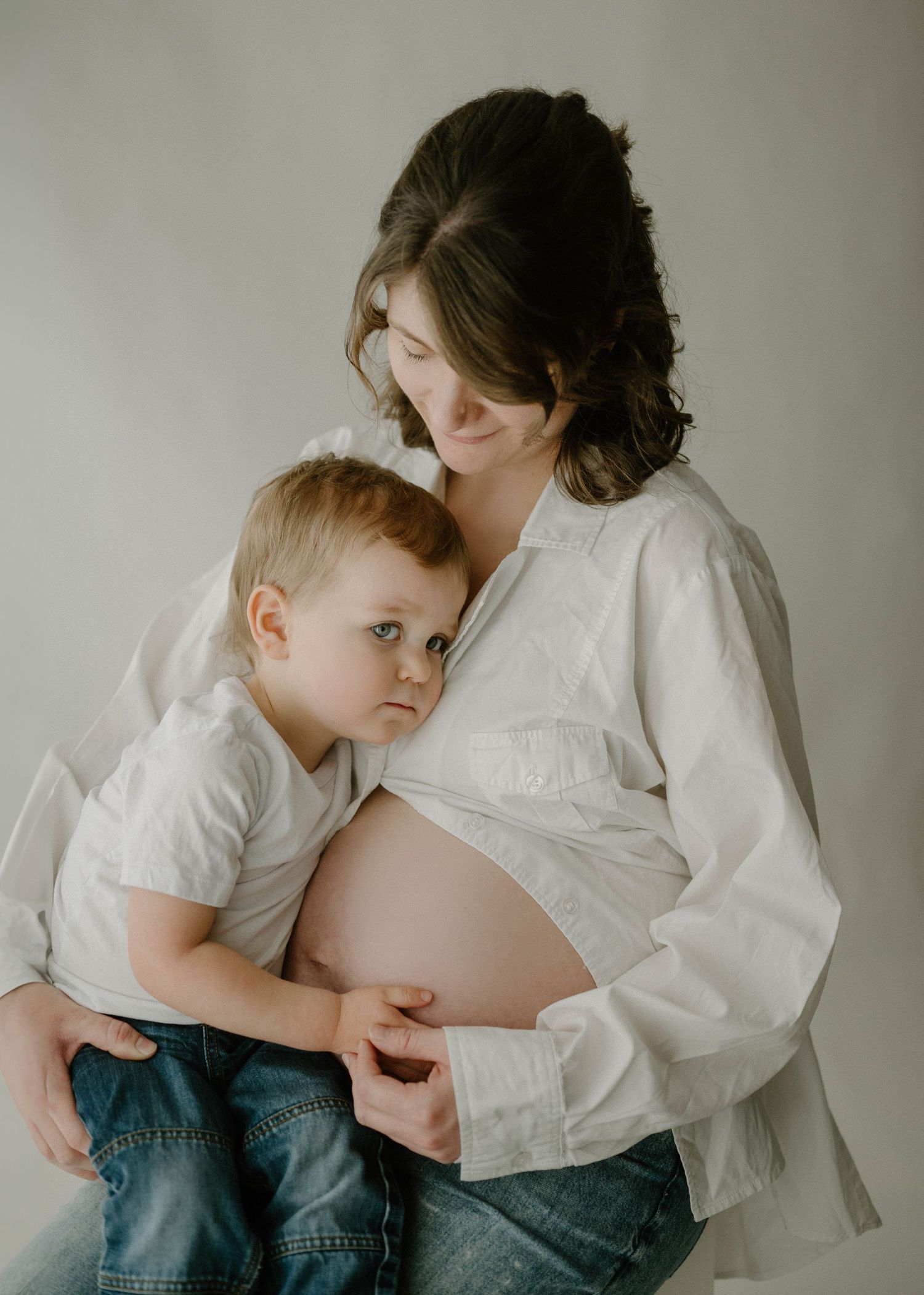 Pregnant woman in white shirt holds toddler boy who rests against her bare baby bump in studio.