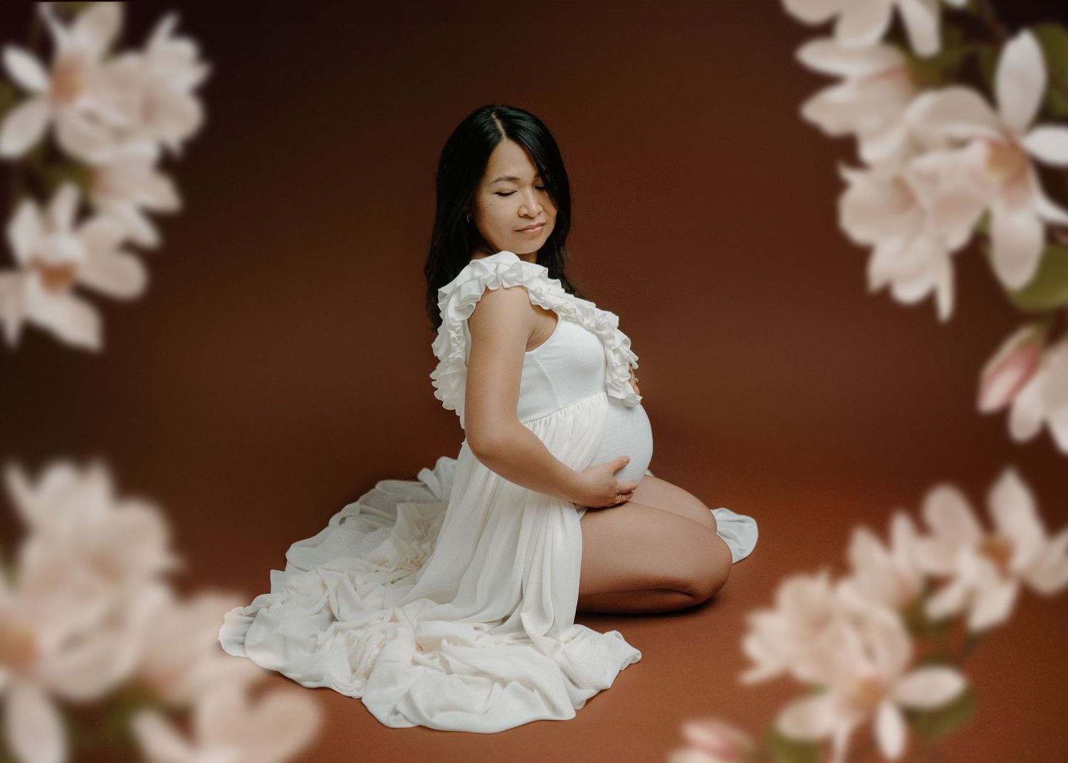 Pregnant woman in white ruffled dress kneeling surrounded by soft pink flowers on brown background.