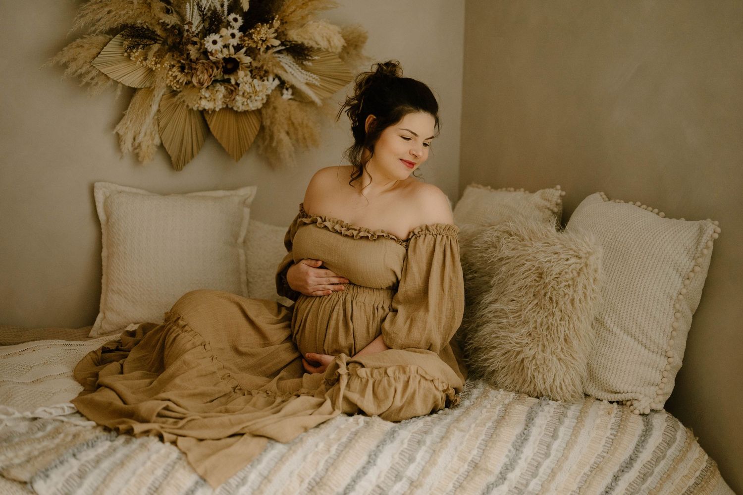 Expecting mother in tan off-shoulder dress sitting on bed with pillows in bohemian styled room.