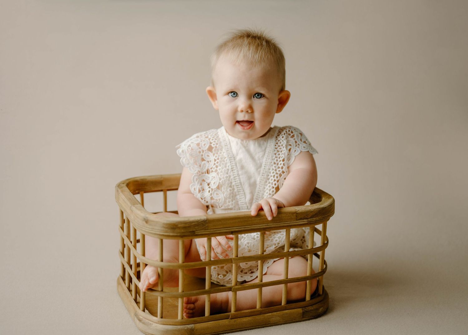 Baby girl in white lace romper sitting in a wicker basket during a studio milestone photo session.