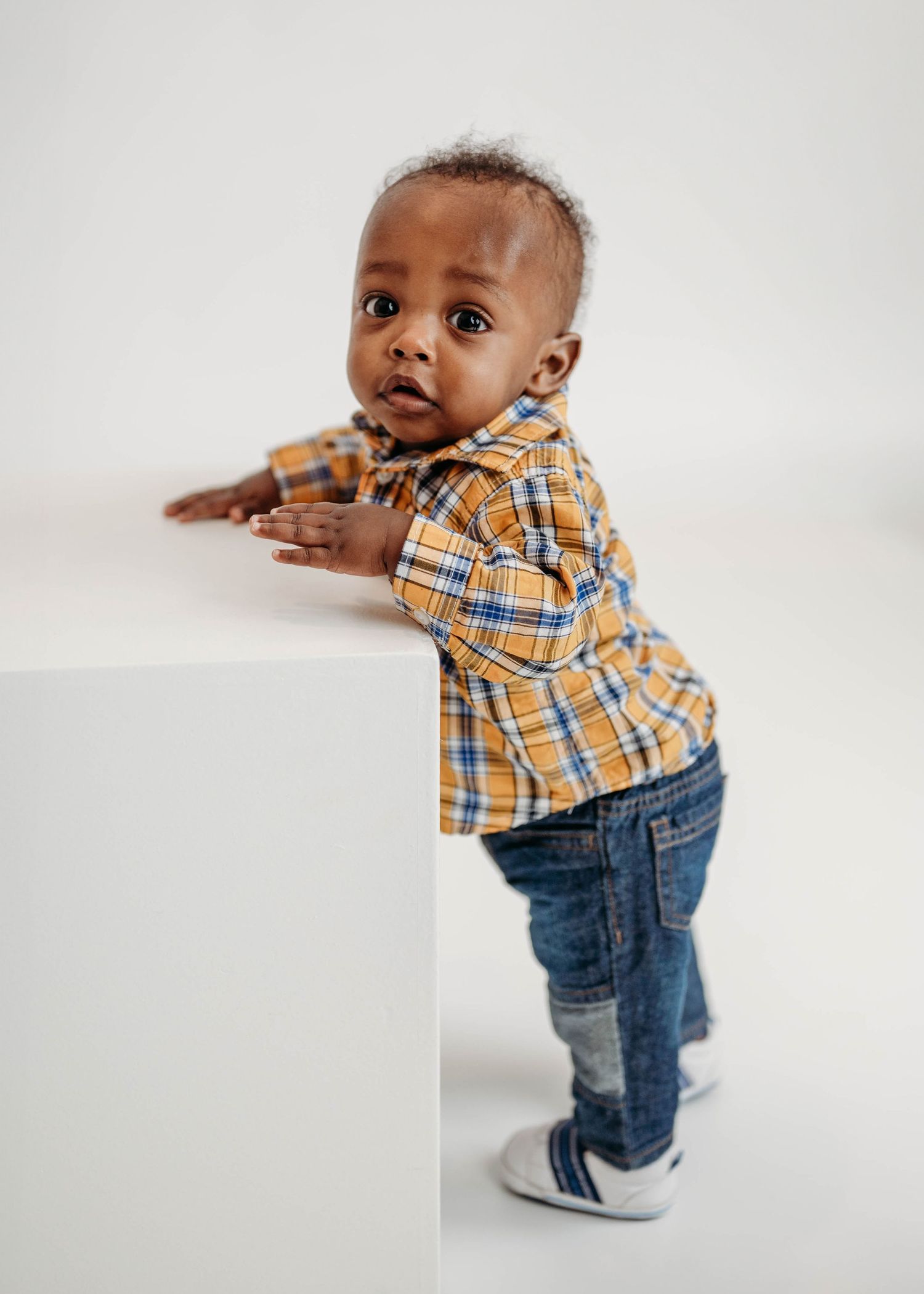 Black toddler boy in a yellow plaid shirt and jeans standing and leaning against a white surface.