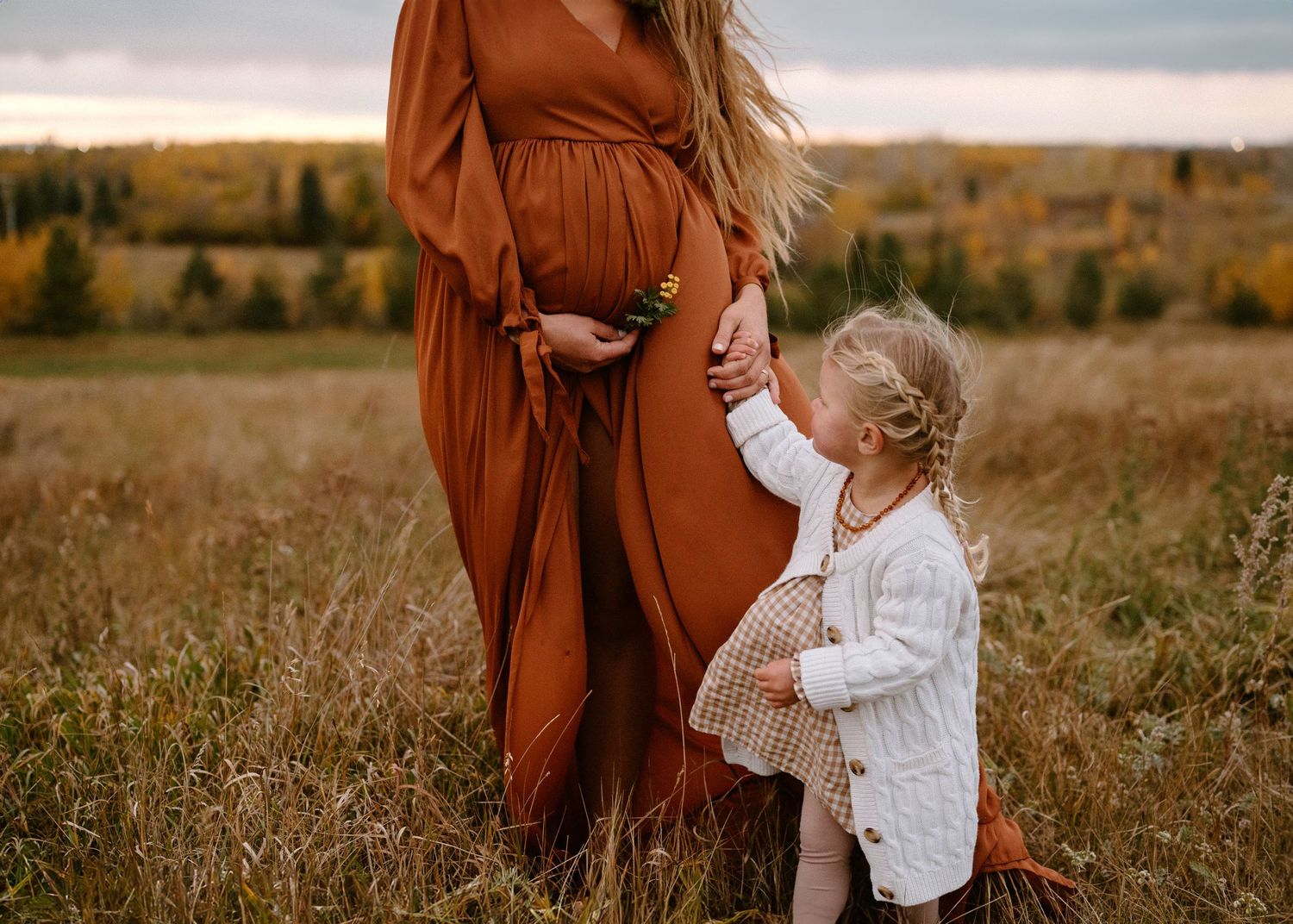 Toddler girl touching pregnant mothers belly in russet maternity dress in fall meadow.