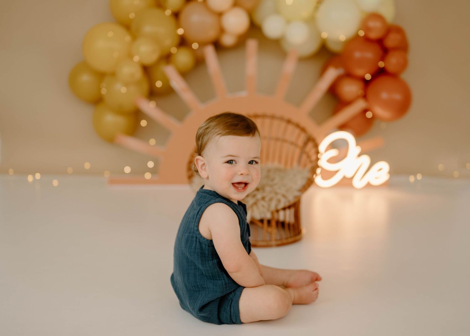 Smiling baby boy in teal romper sitting on floor with boho sun balloon arch birthday backdrop.