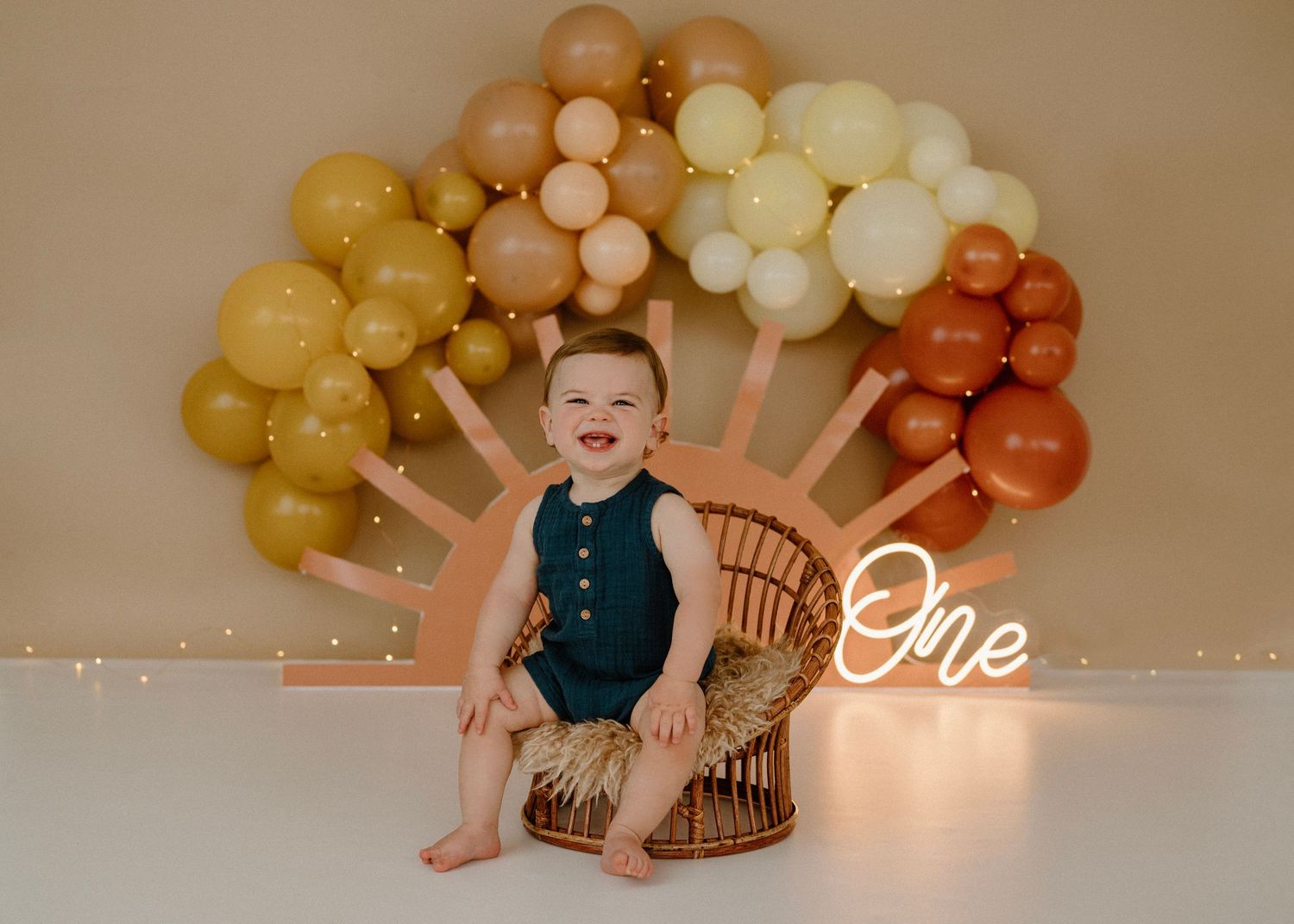 Smiling baby boy sitting in wicker chair at one year cake smash with rainbow balloon backdrop.