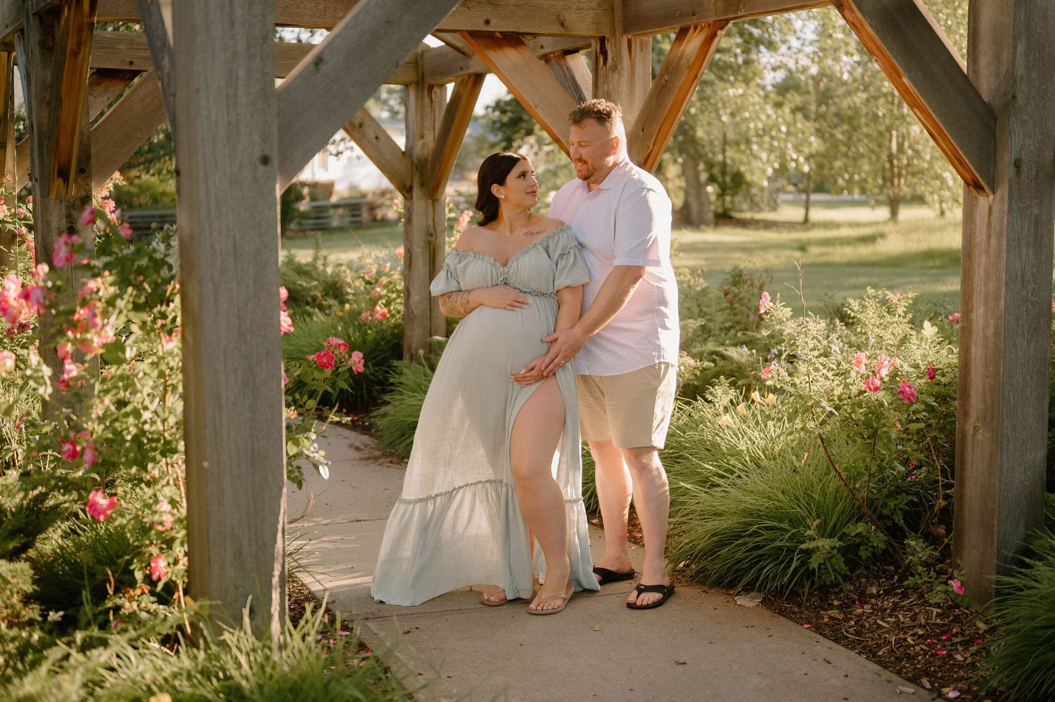 Pregnant couple posing under wooden arbor surrounded by blooming flowers in a sunny garden during maternity photo session.