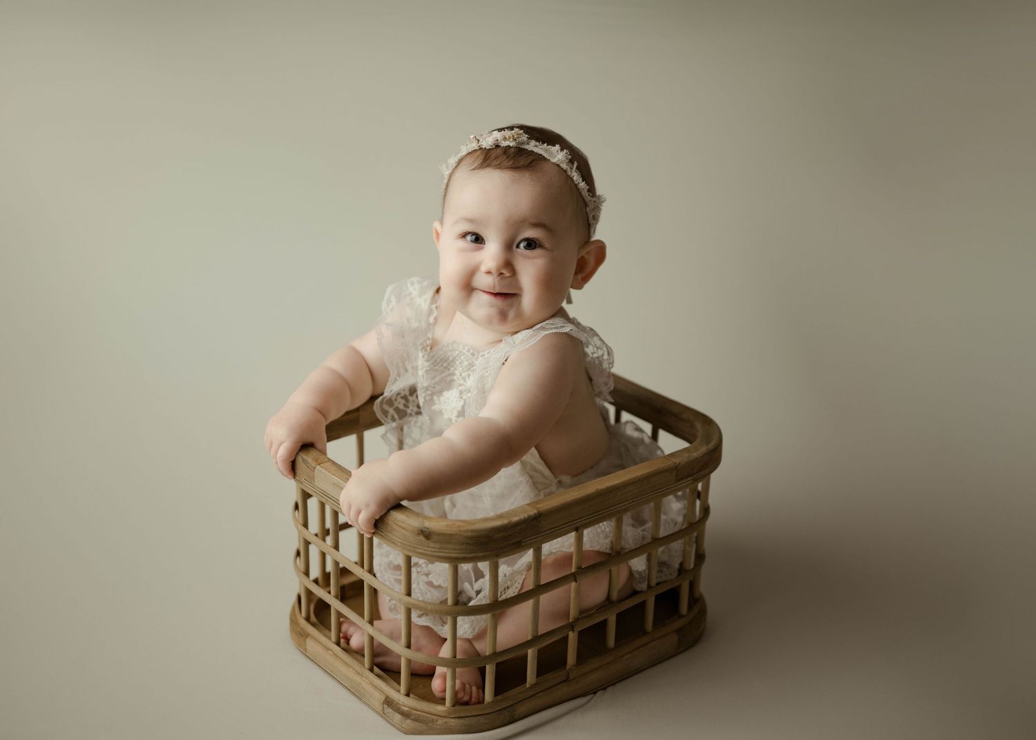 Cheerful baby girl in white lace dress and headband smiling inside a rustic wooden basket prop.