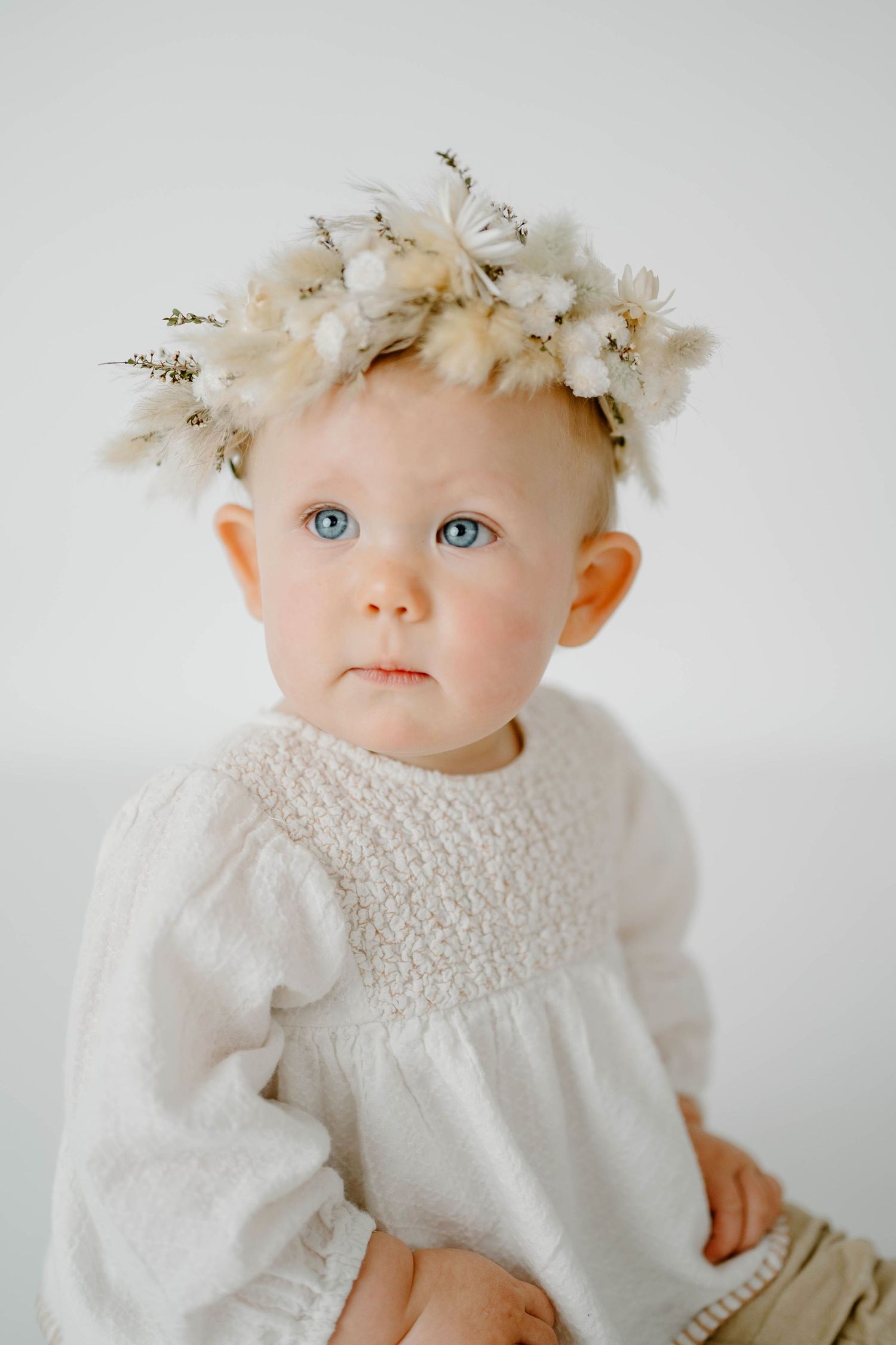 Sweet baby girl with blue eyes wearing white floral crown and smocked white dress in studio.