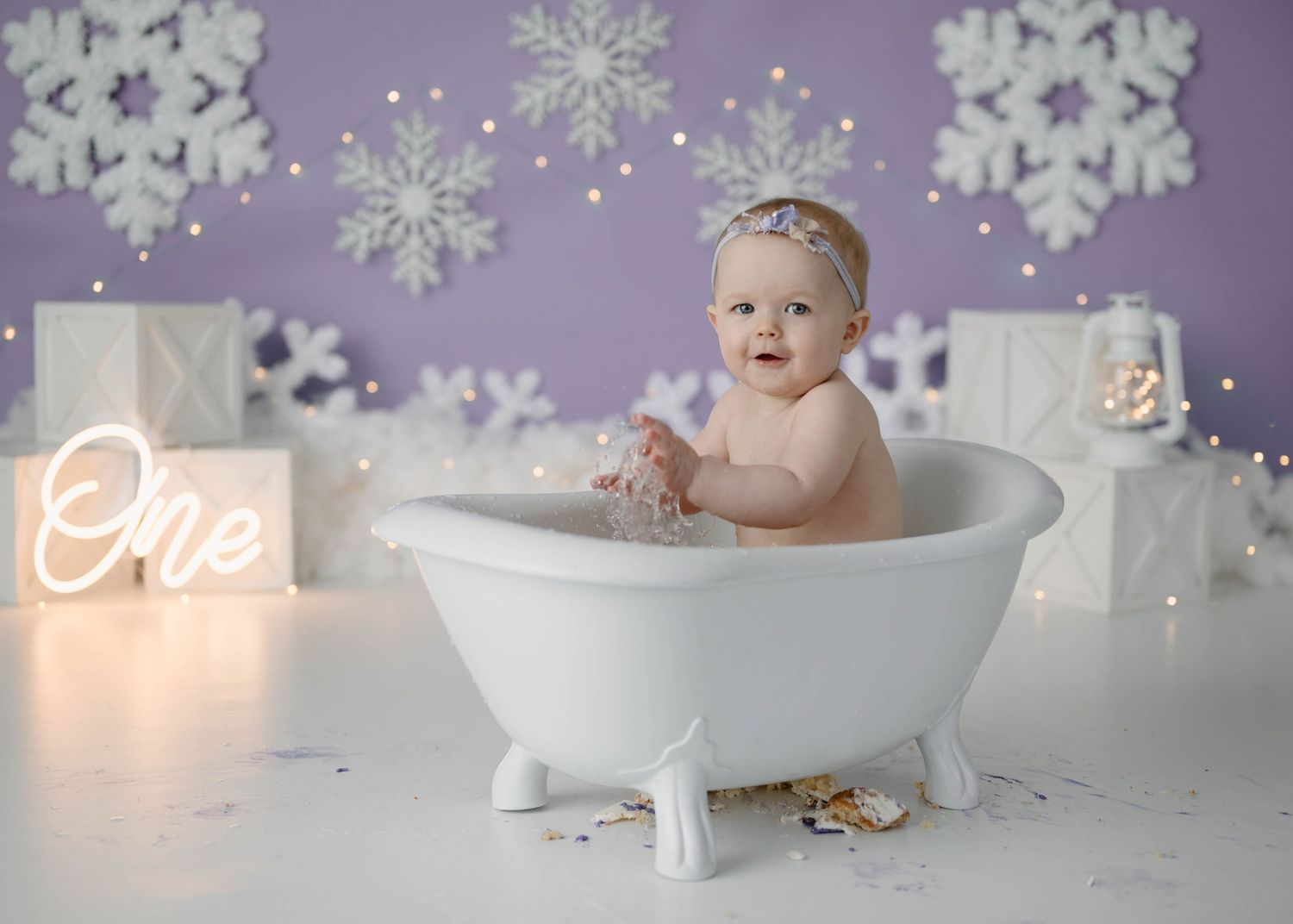 Happy baby sits in small white bathtub surrounded by snowflake decorations and One neon sign birthday setup.