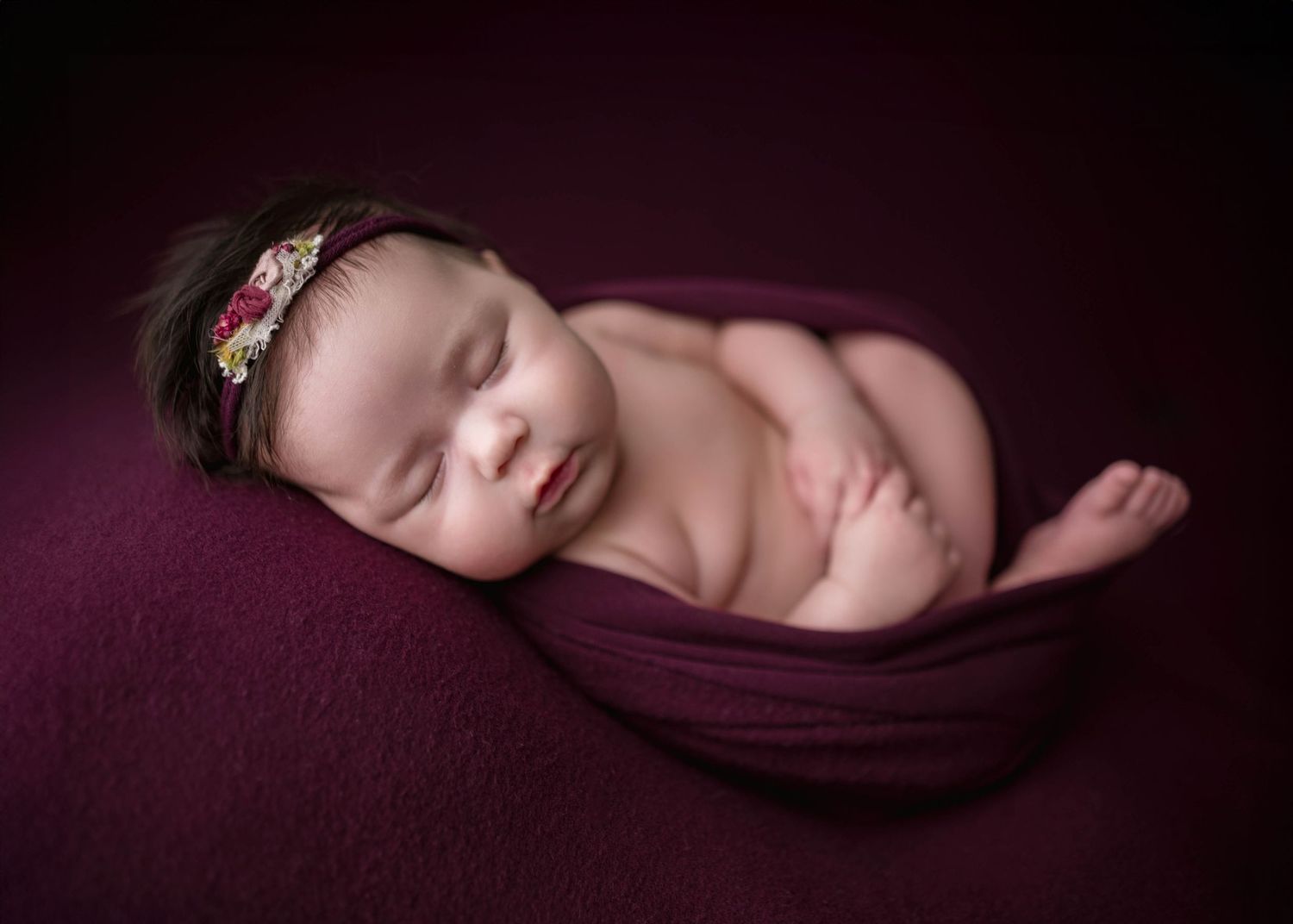 Newborn baby girl sleeping peacefully wrapped in burgundy fabric with floral headband on matching background.