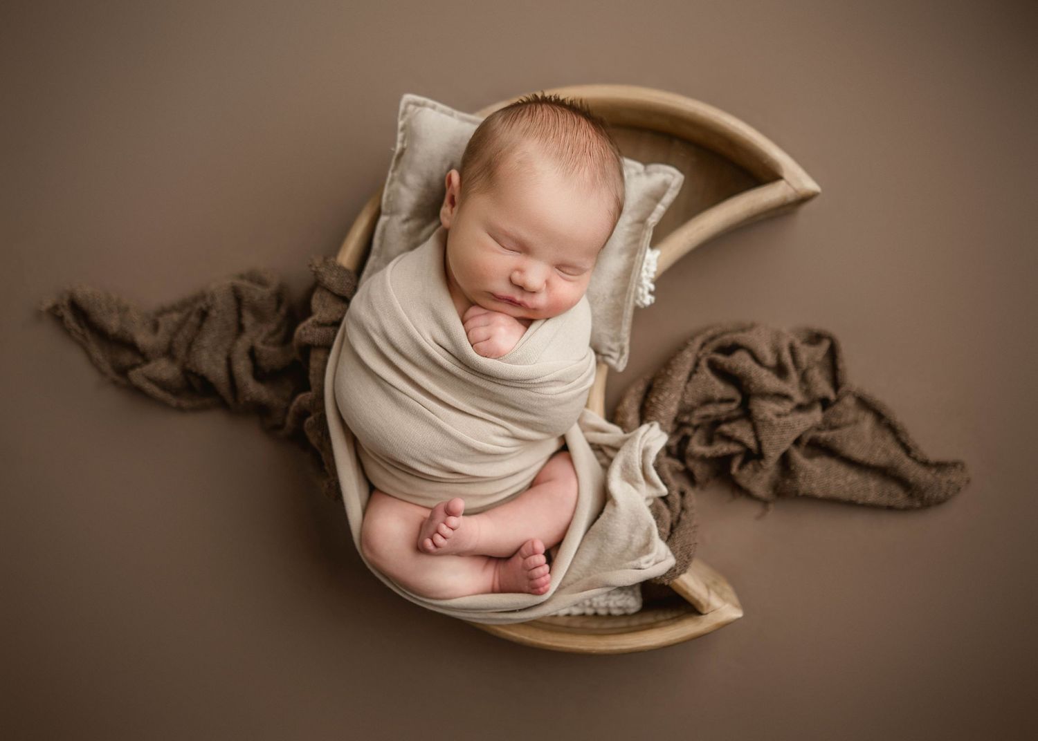 Newborn baby swaddled in cream wrap sleeping peacefully in a wooden moon-shaped bowl prop on brown backdrop.