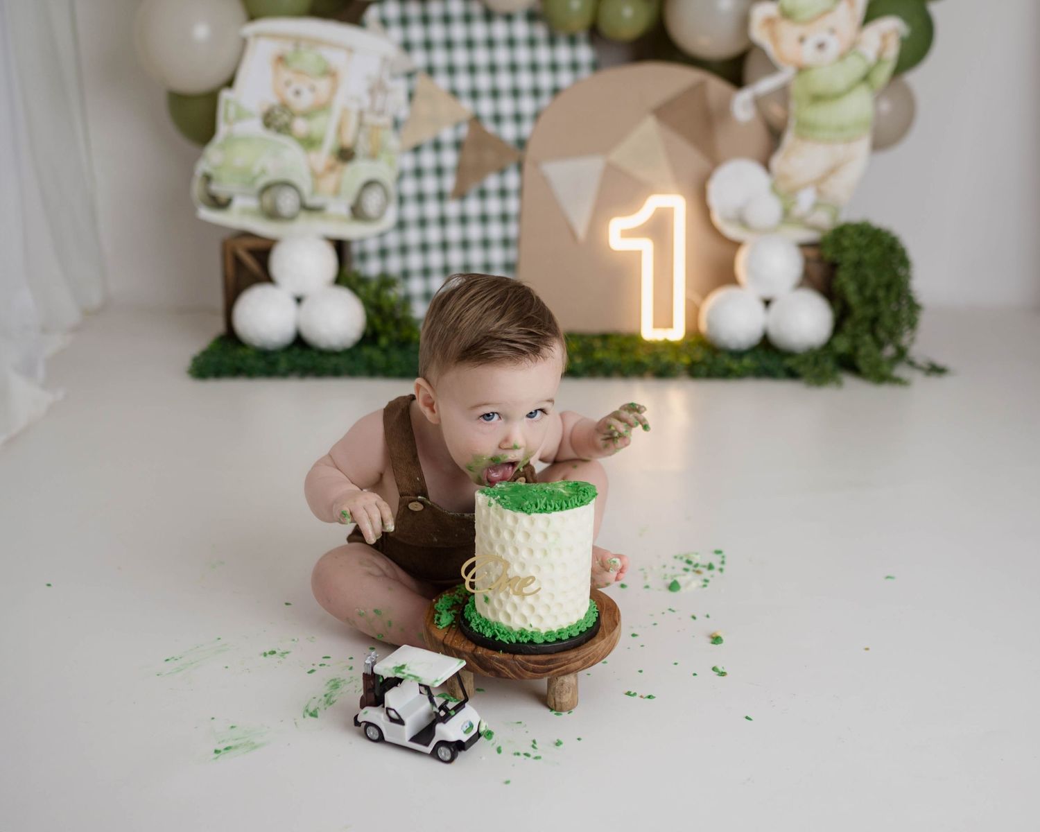 Baby boy smashes golf-themed first birthday cake with green frosting, surrounded by balloons and a number one light.