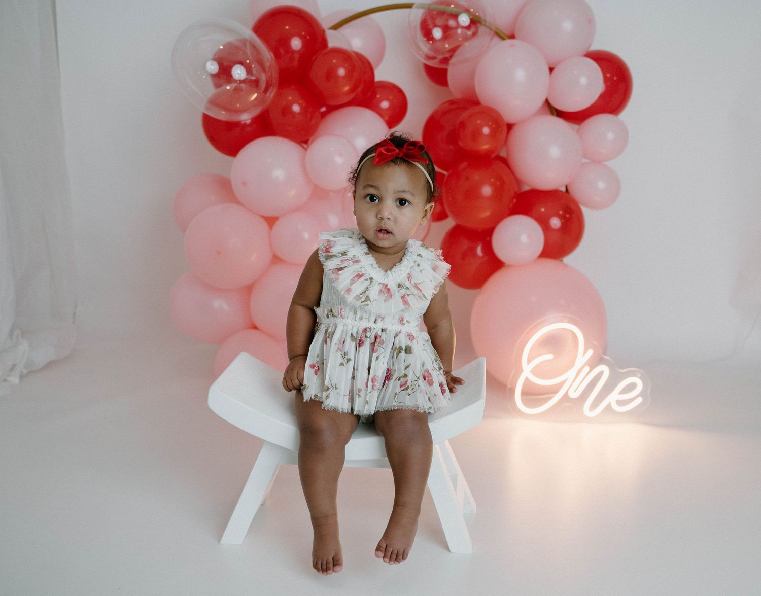 Baby girl in floral romper stands on white chair with red and pink balloon arch and neon One sign.