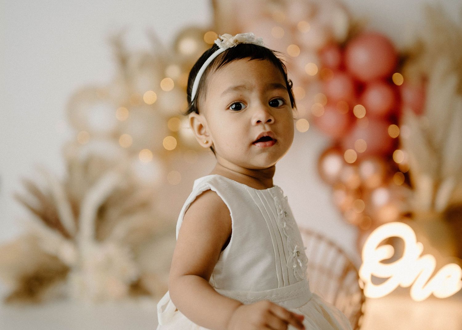 Adorable baby girl with headband poses in front of glowing balloon arch at first birthday cake smash session.