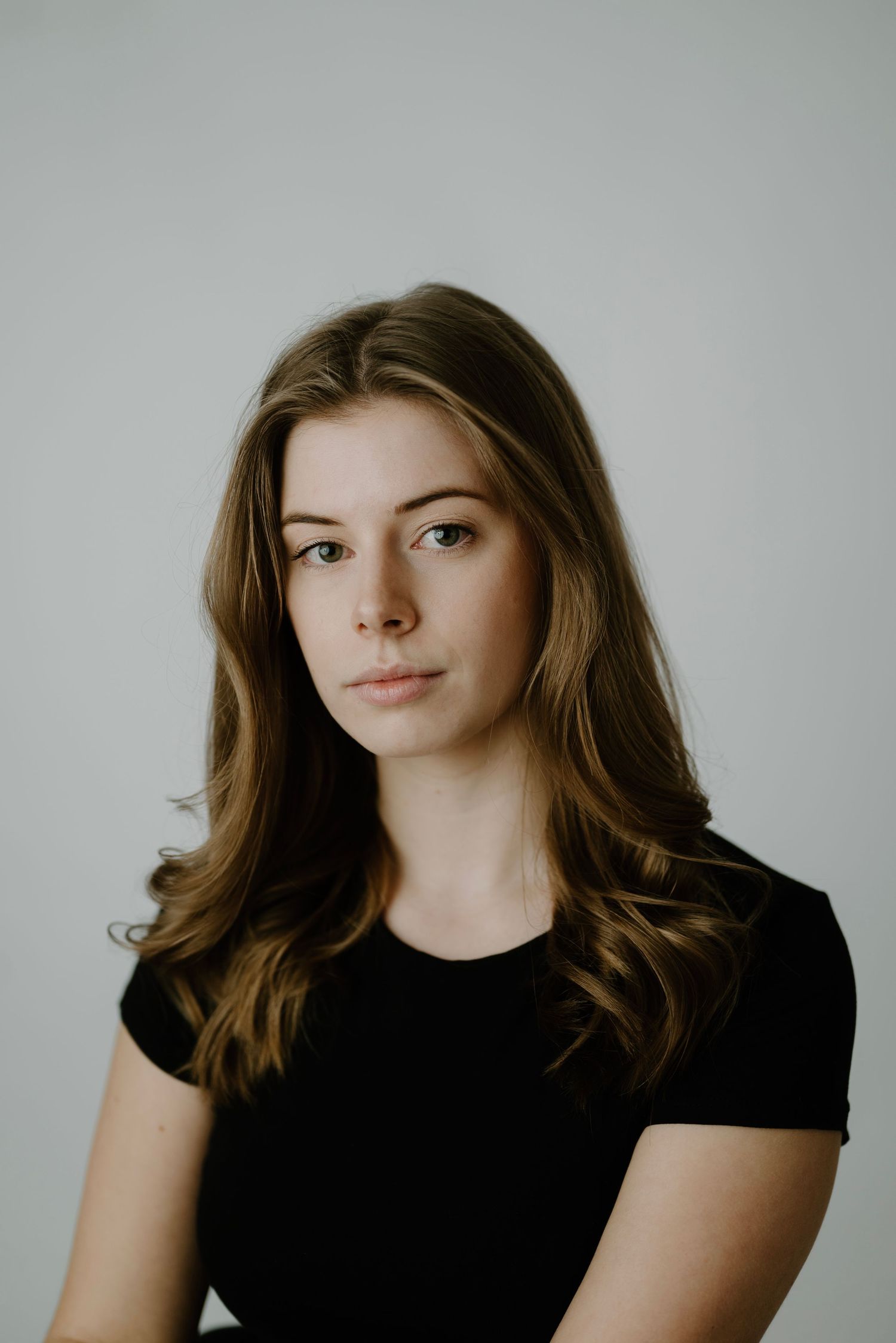Young woman with long brown hair wearing a black t-shirt poses for a professional headshot against a gray background.