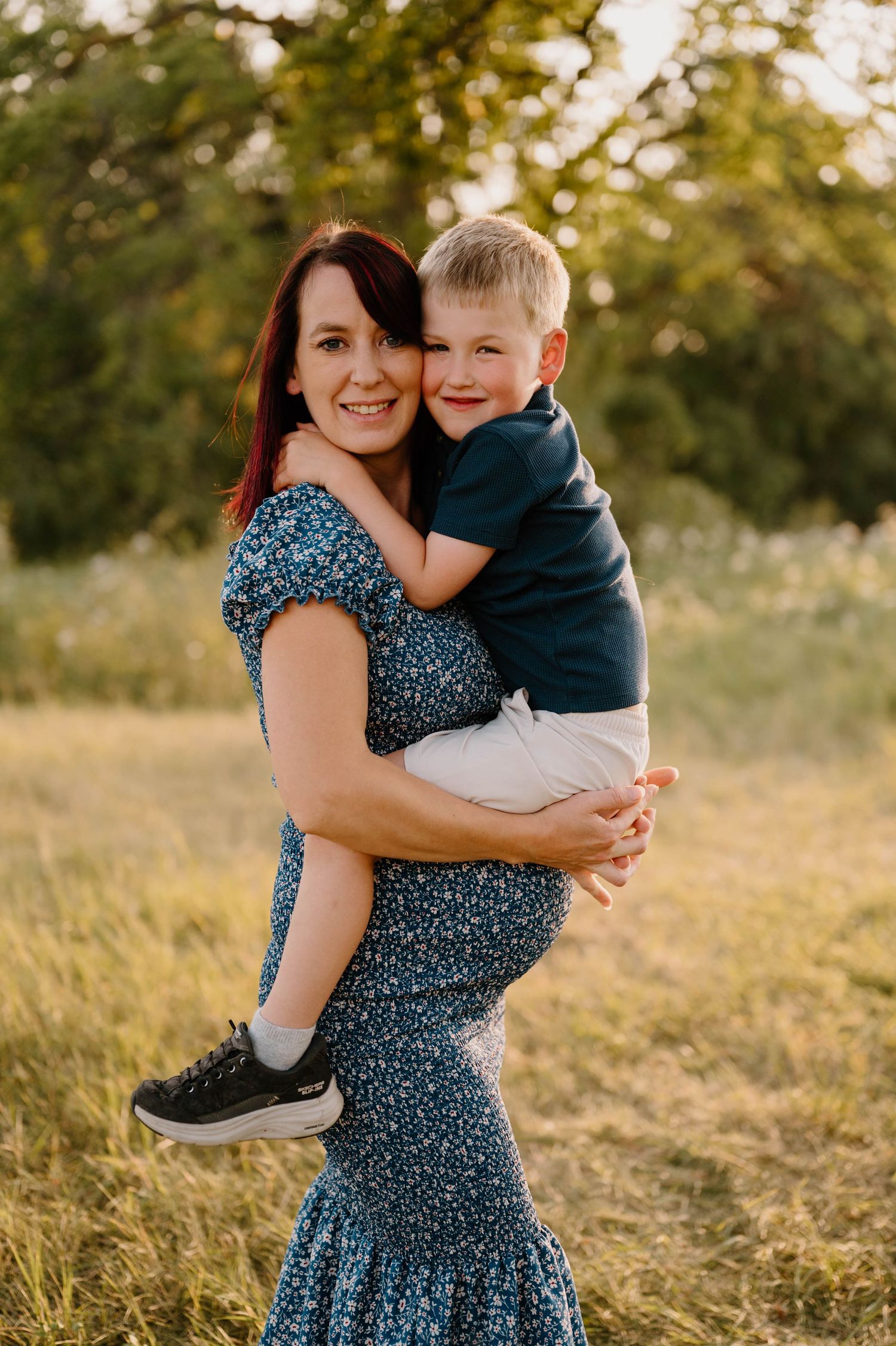 Smiling redhead pregnant mother holding young blond son in sunny green meadow at sunset.