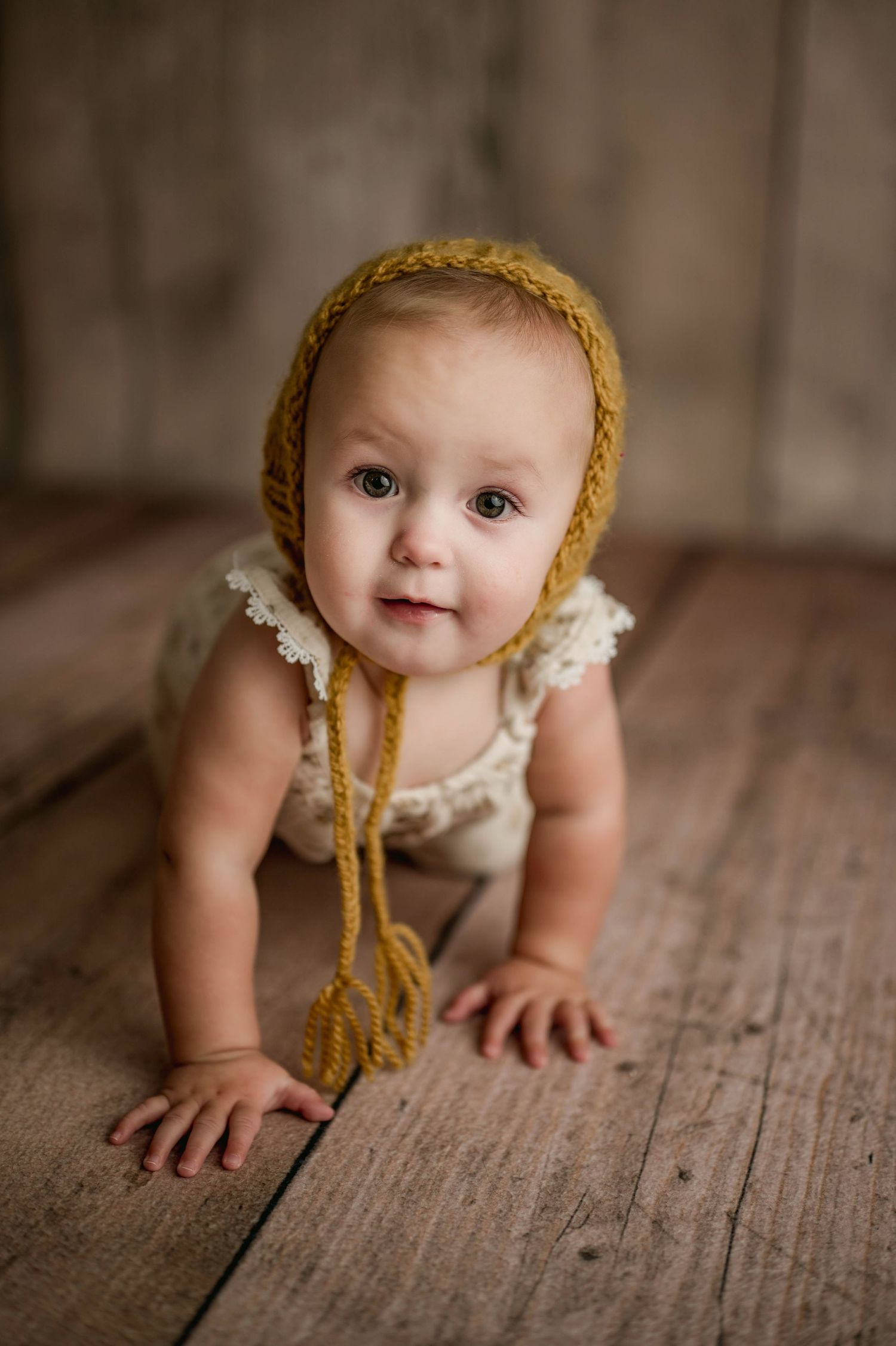 Baby in cream romper and mustard yellow knit bonnet crawls on rustic wood floor in studio.