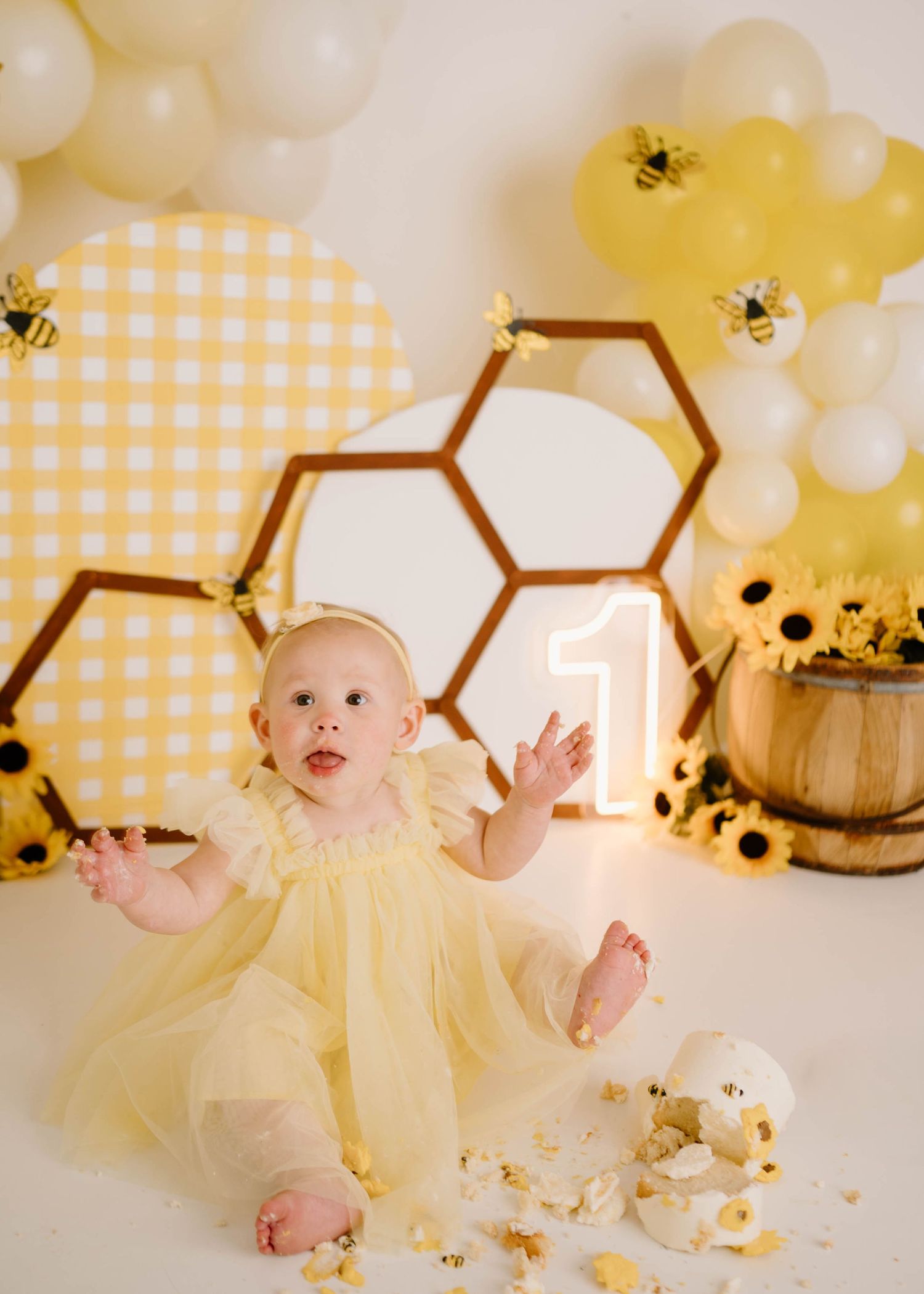 Baby girl in yellow tutu dress at a bee-themed first birthday cake smash with honeycomb decor and balloons.
