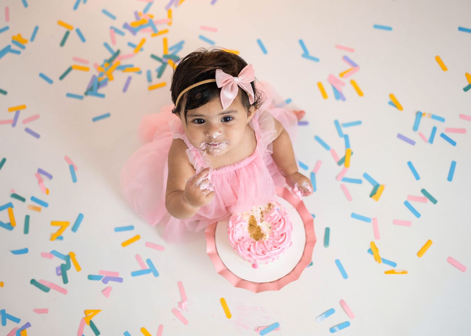 Overhead view of baby in pink tutu with smash cake surrounded by colorful sprinkles on white floor.