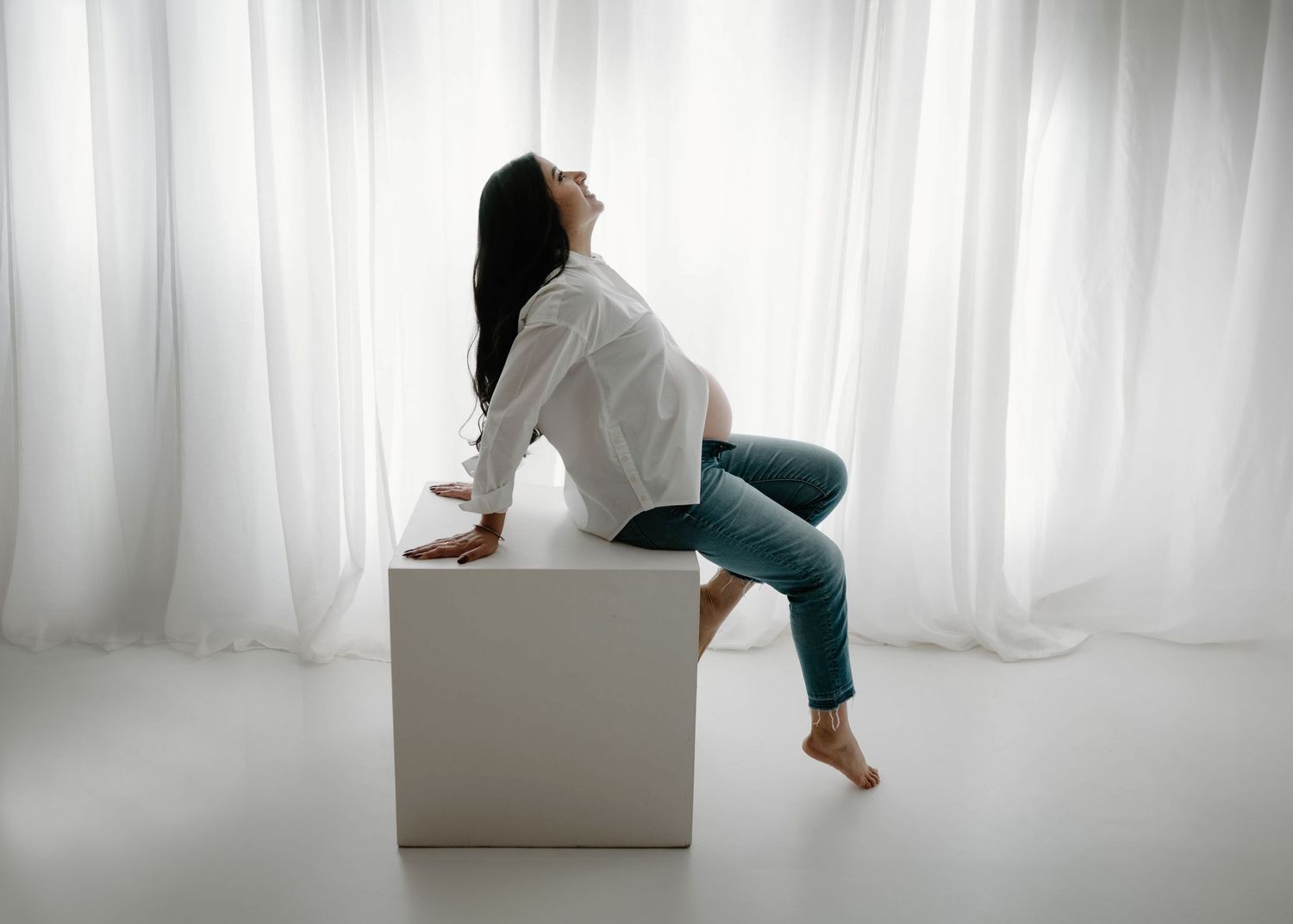 A woman in white shirt and jeans sits on a white cube, leaning back gracefully in a bright studio.