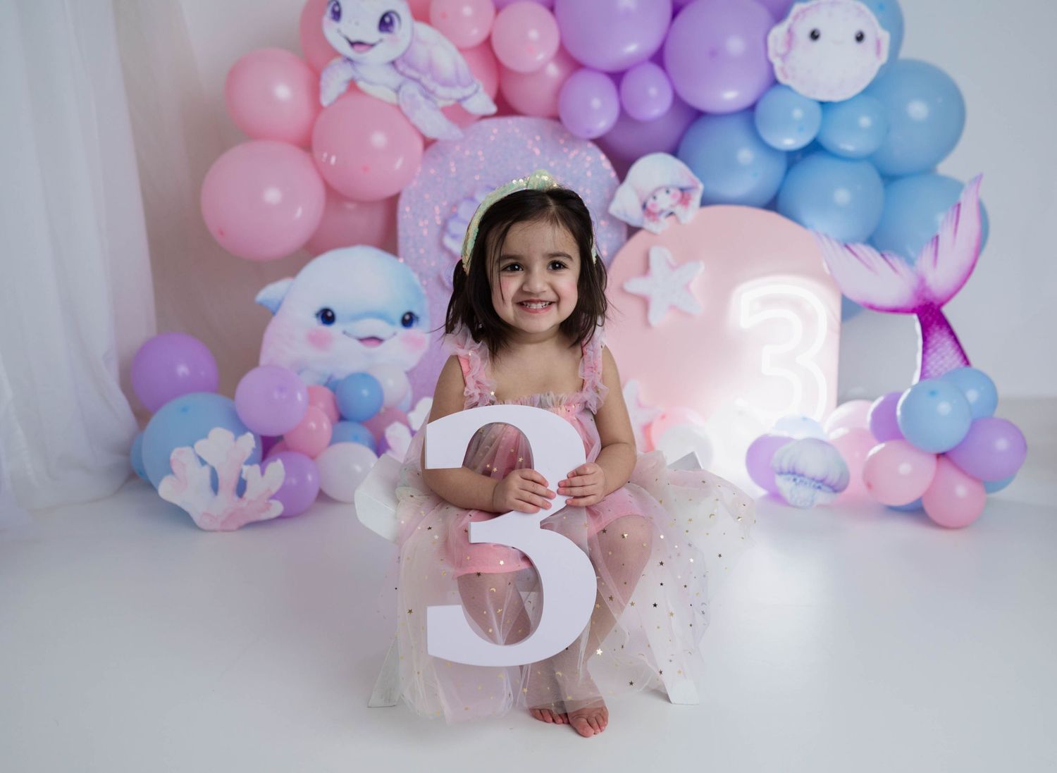 Little girl in pink dress holding number three at mermaid themed third birthday party setup.