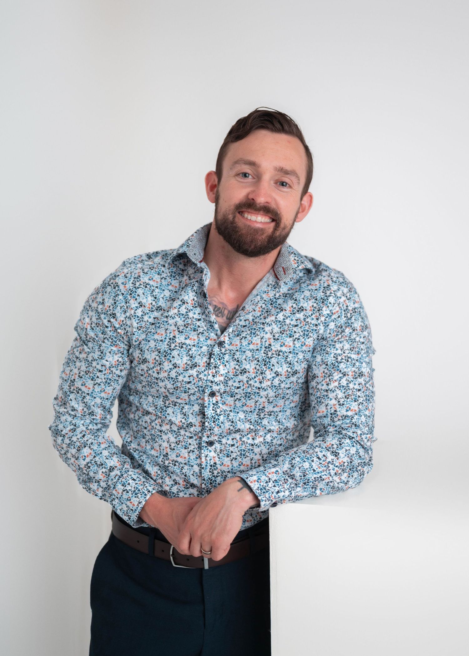 Smiling bearded man in a blue floral button-up shirt posing confidently against a light gray background.