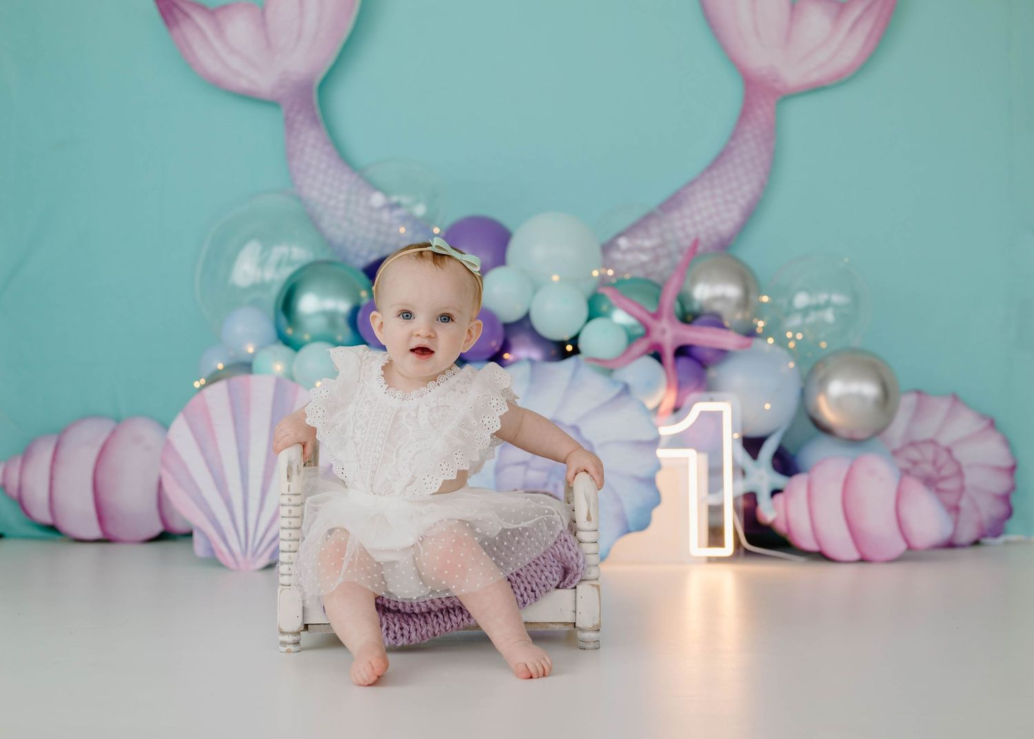 Baby girl in white dress sitting on chair at mermaid-themed first birthday party with shells.