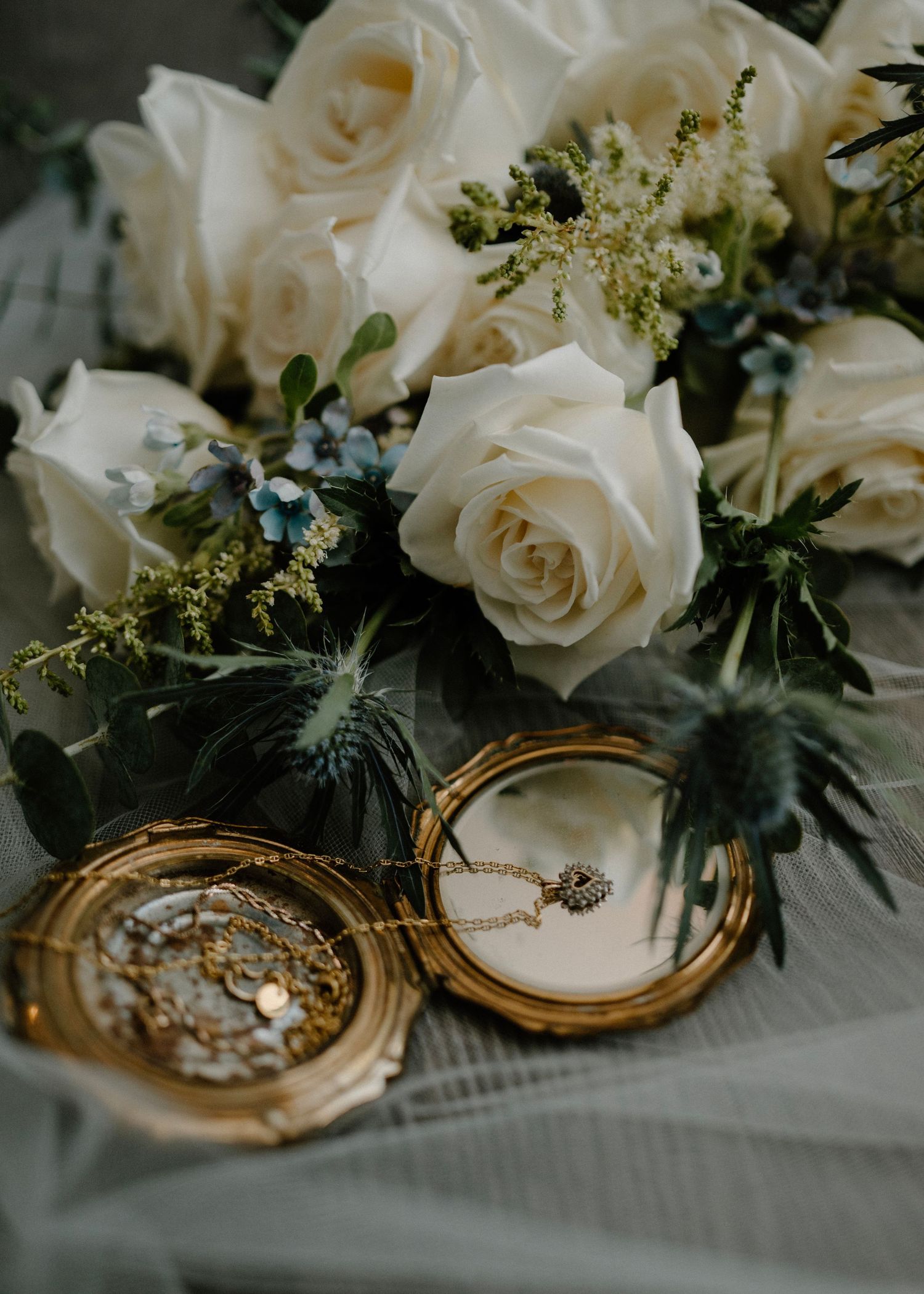 Gold vintage locket with necklace displayed beside a bridal bouquet of white roses and blue thistle.